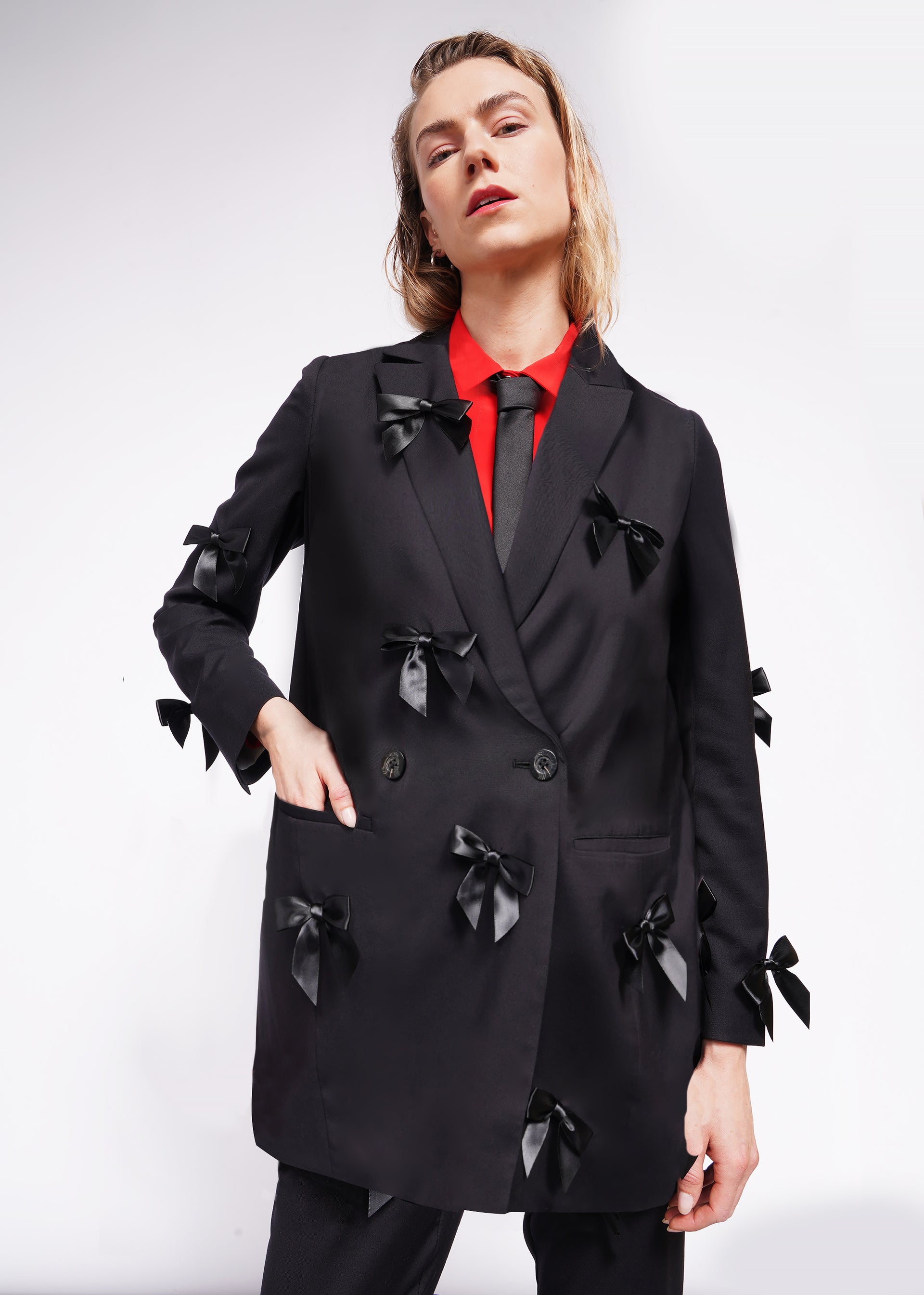 S= A person is wearing The Empower Bows Double Breasted Blazer, tailored and oversized with multiple black bows, paired with a red shirt and a black tie. They stand with one hand in their pocket against a plain white background.