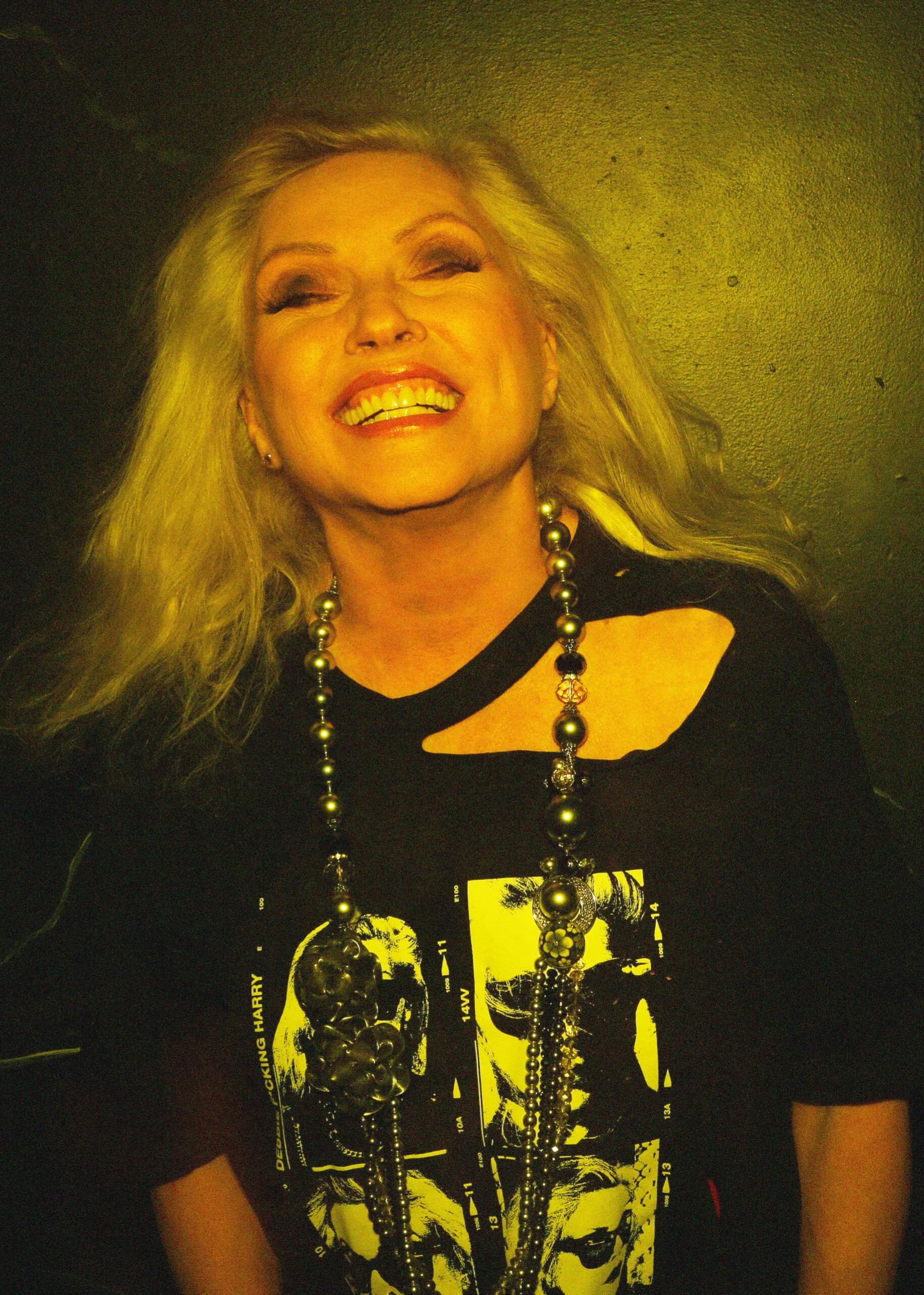 Person with long blonde hair wearing a black shirt with graphic design, against a dark background.