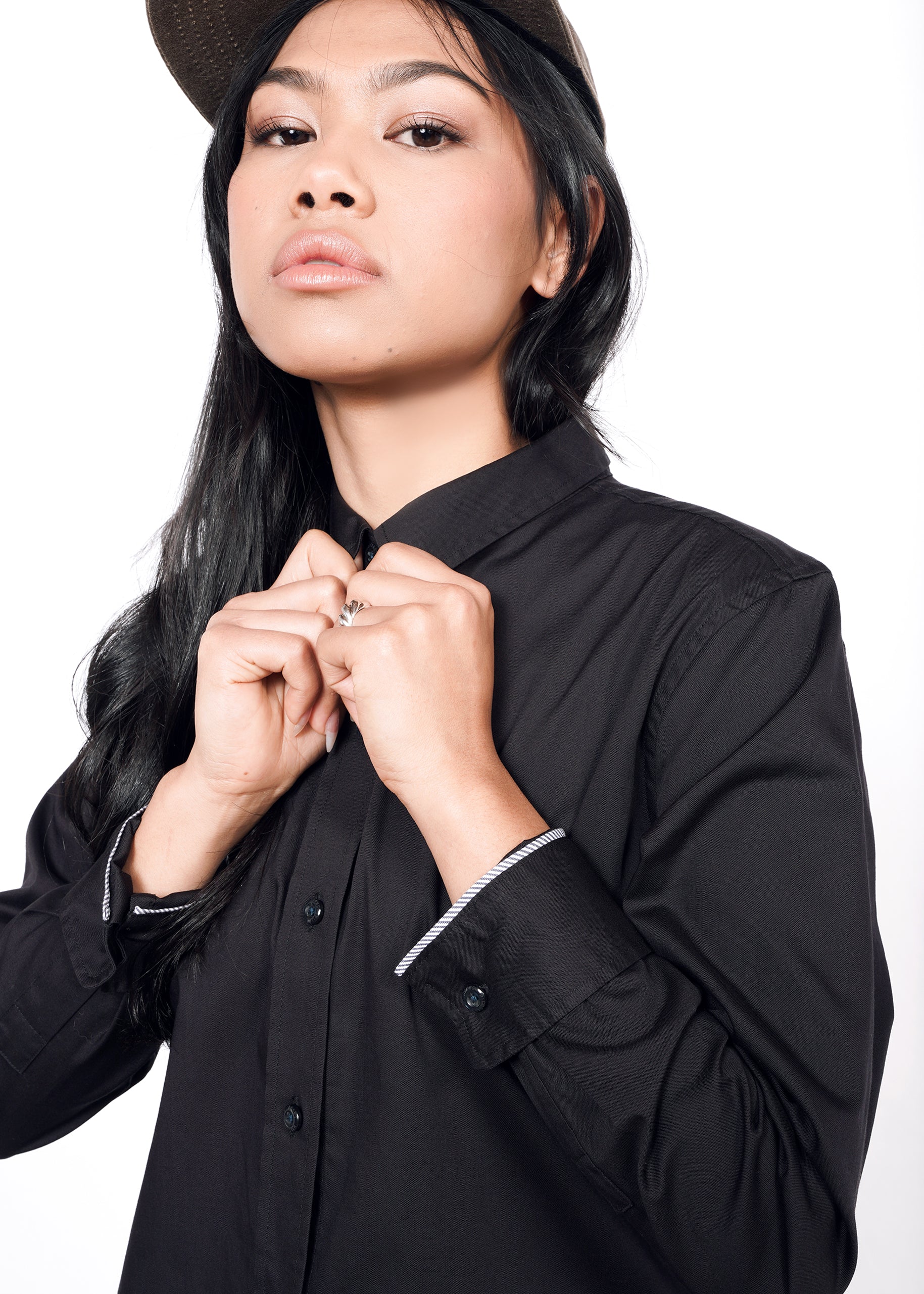 S=A person with long black hair confidently adjusts their collar, wearing The Essential Long Sleeve Oxford Button Up and a cap, against a white background.