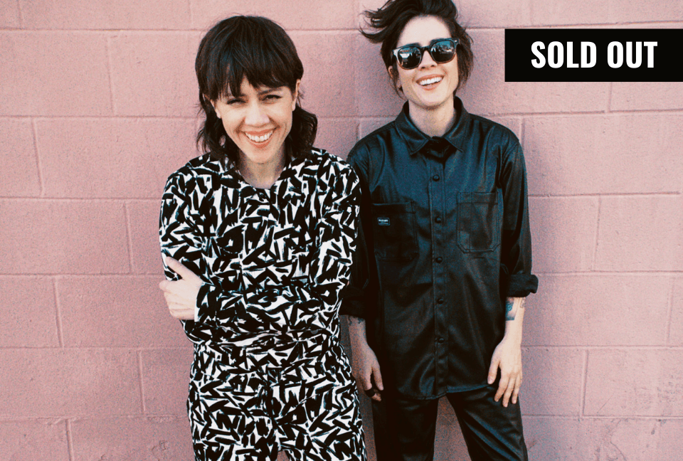 Tegan and Sara look at camera standing in front of pink brick background. One is wearing a black and white print Wildfang coverall and the other is wearing a matching faux leather top and pants. A banner in the top right reads SOLD OUT.