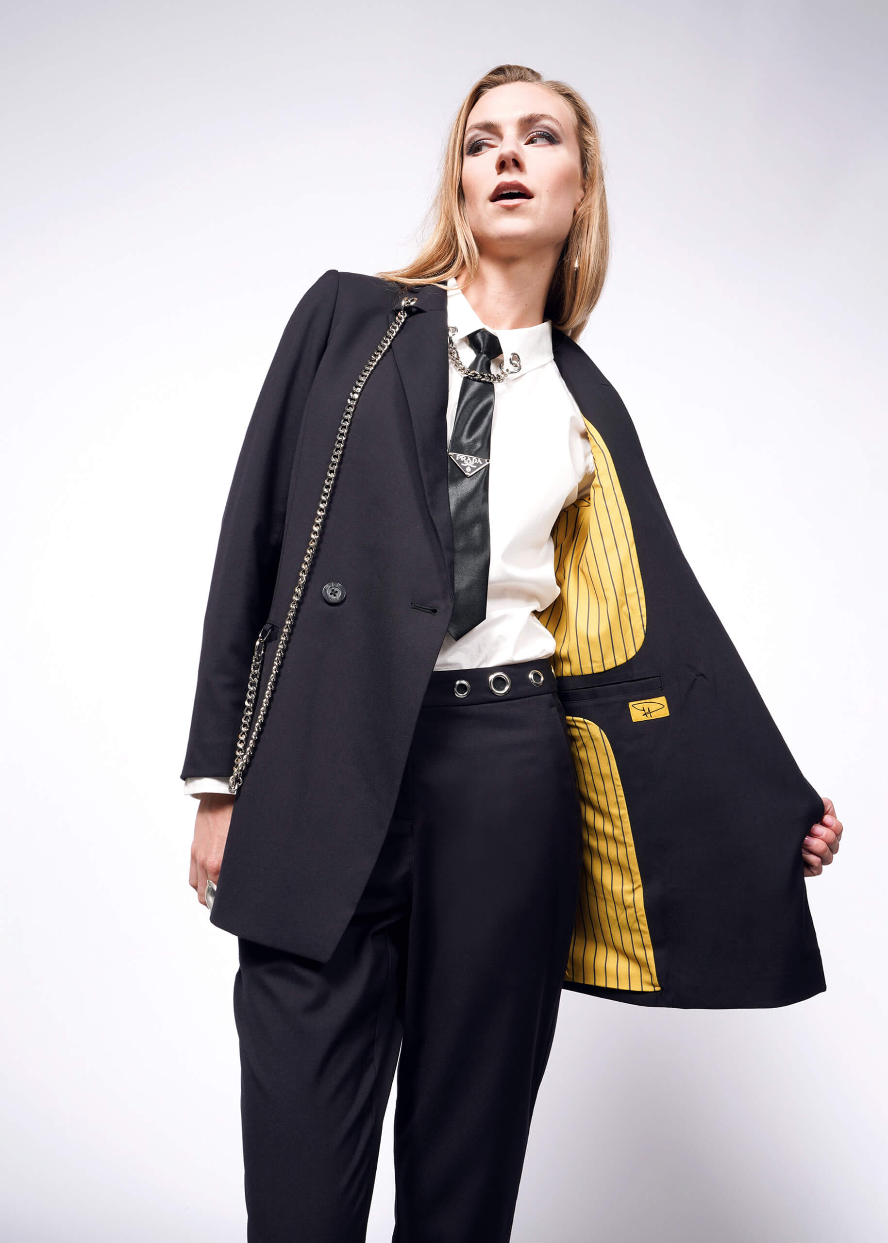 S=Dressed in a stylish black suit featuring the Debbie Harry X Wildfang Slim Crop Pant, a person poses confidently. They reveal the blazers yellow-striped lining over a white shirt and black tie, accentuated by chain details, embodying a punk aesthetic against a plain white background.