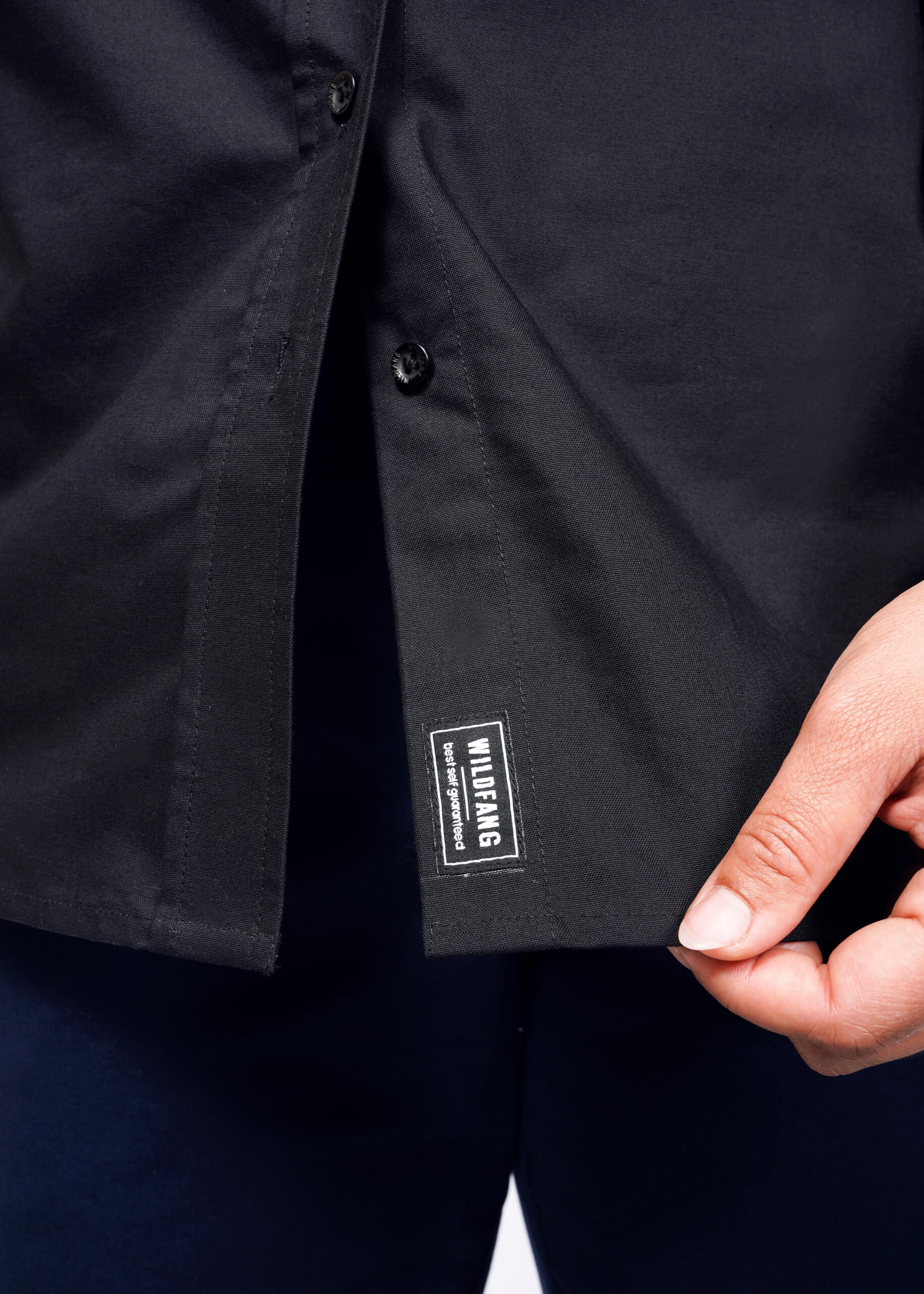 1X=A person lifts the hem of The Essential Long Sleeve Oxford Button Up, a black, tailored-fit shirt, exposing a small WILDFANG label in white. This 100% cotton shirt is unbuttoned at the bottom and elegantly paired with dark blue pants.