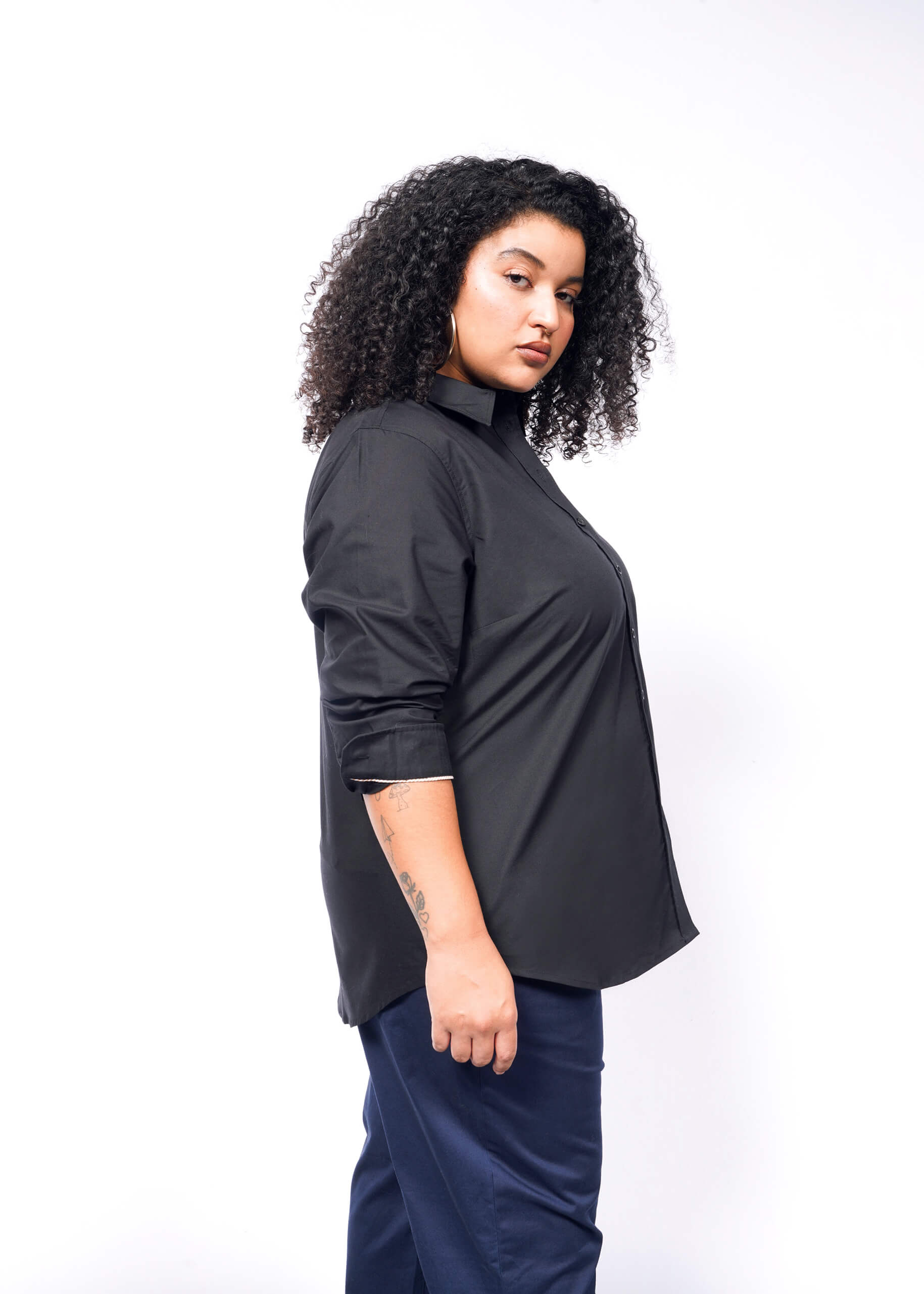 1X=Against a plain white background, a person with curly hair stands in profile. They wear The Essential Long Sleeve Oxford Button Up in black, with rolled-up sleeves and perfectly tailored blue pants, exuding a confident expression.