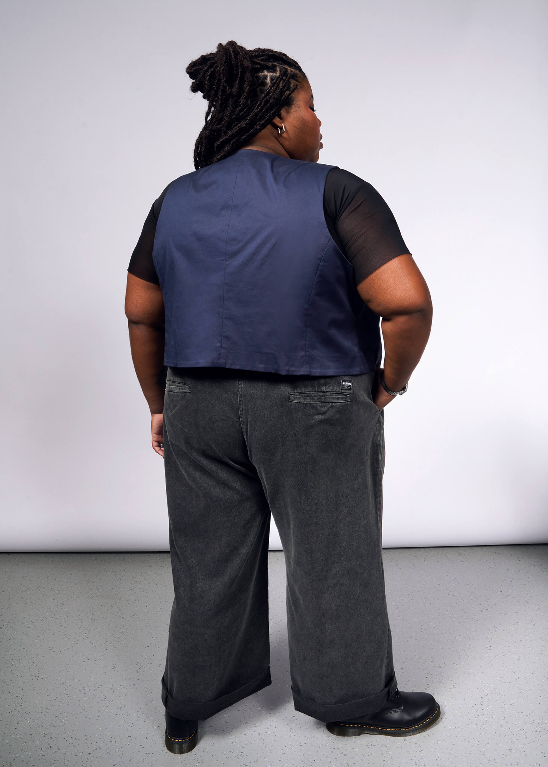1X=A person with long braided hair faces away from the camera, wearing a black t-shirt, The Essential Vest for layering, gray wide-leg pants, and black boots. They stand on a light gray floor against a white background.