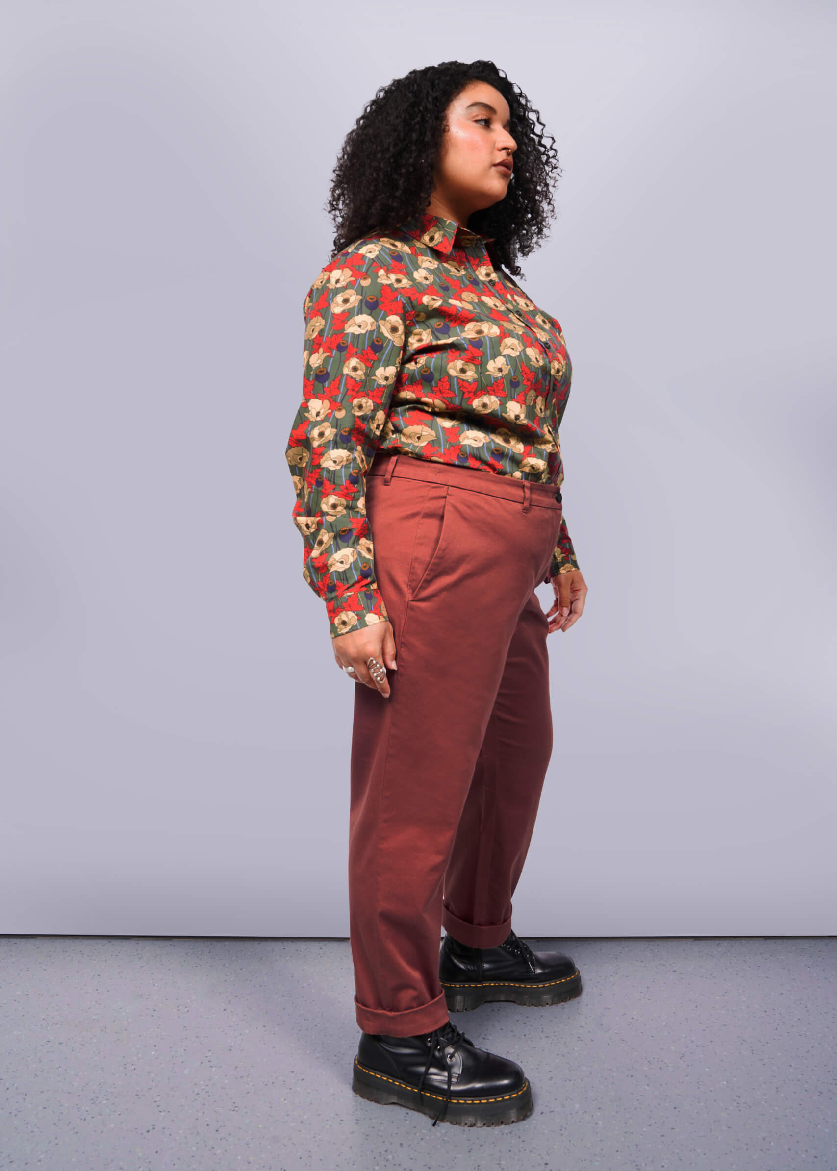 1X=With their curly hair, a person confidently wears The Essential Trouser featuring a relaxed mid-rise fit. A colorful floral blouse tops the wardrobe staple, paired with black platform shoes for an edgy touch. The plain white background highlights their stylish ensemble.
