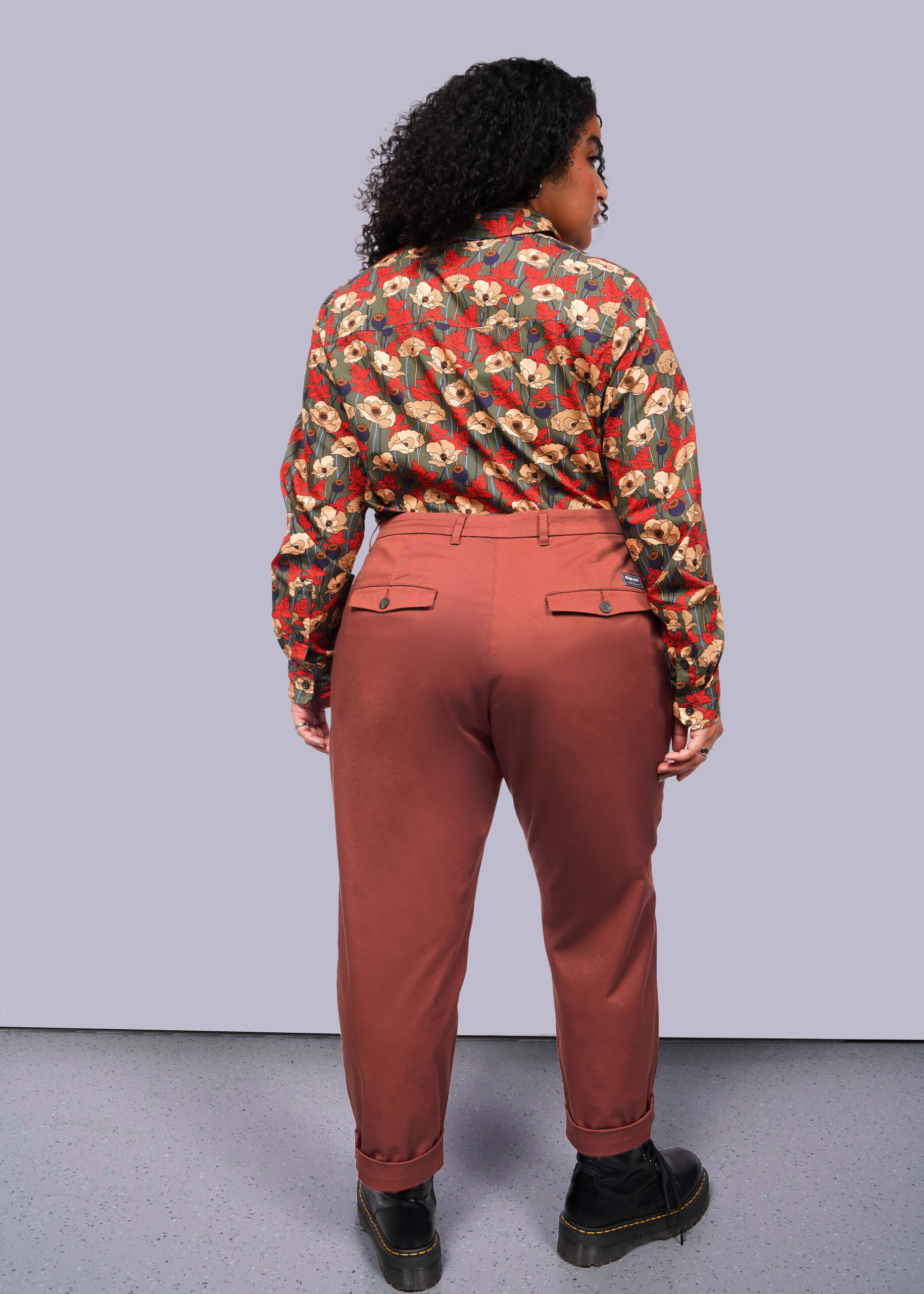 1X=A person with curly hair stands with their back to the camera, showcasing The Essential Trouser in a classic rust color and relaxed mid-rise fit, paired with a floral shirt. Black shoes complete this timeless ensemble against a plain white backdrop.
