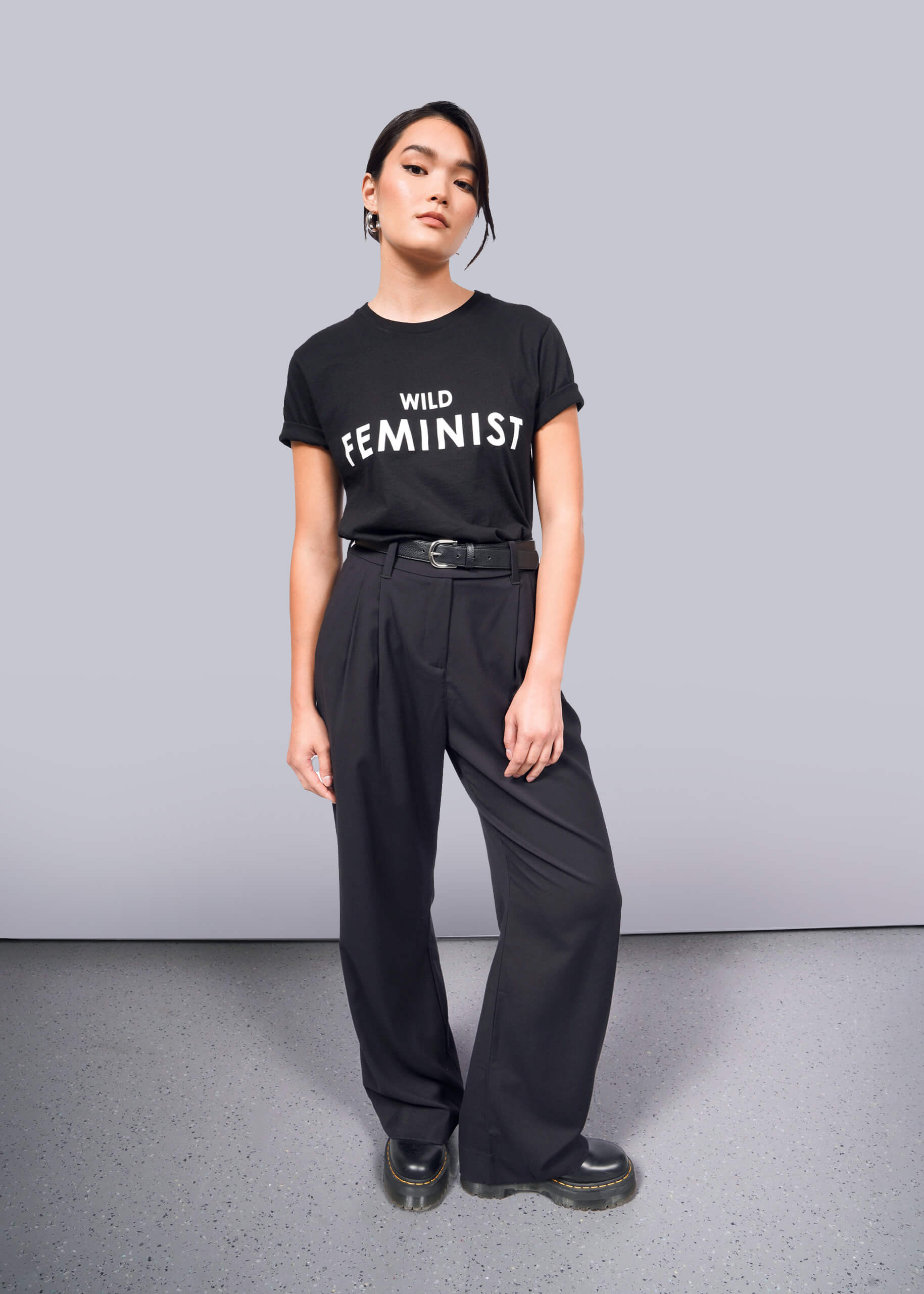 S=Against a white background, someone stands confidently in The Wild Feminist® Tee, a 100% cotton black T-shirt. They pair it with belted black pants and chunky-soled shoes, with hands relaxed at their sides.