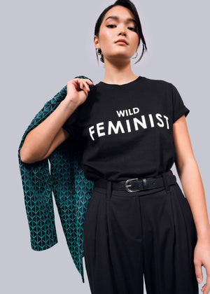 S=A woman confidently poses in The Wild Feminist® Tee, a black unisex fit T-shirt with WILD FEMINIST in bold white letters, made from pre-shrunk 100% cotton for lasting comfort. She holds a jacket over her shoulder and pairs it with high-waisted black pants against a plain white background.