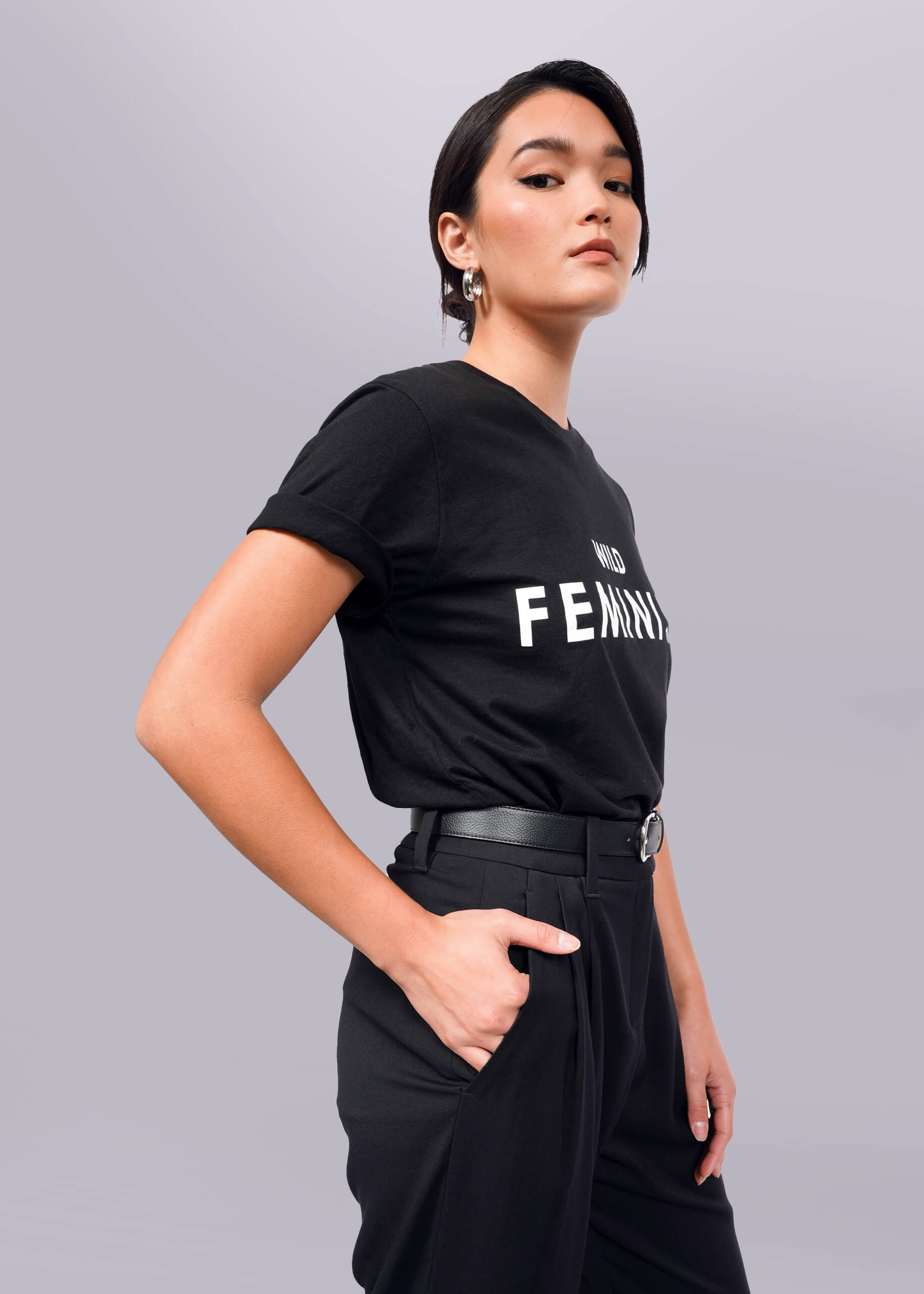 S=Confidently standing against a plain backdrop, a short-haired woman with hoop earrings wears The Wild Feminist® Tee, a unisex black cotton t-shirt with FEMINIST boldly displayed. Her hand in her pocket complements her bold stance.