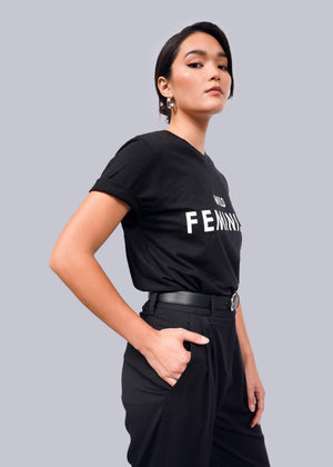 S=Confidently standing against a plain backdrop, a short-haired woman with hoop earrings wears The Wild Feminist® Tee, a unisex black cotton t-shirt with FEMINIST boldly displayed. Her hand in her pocket complements her bold stance.