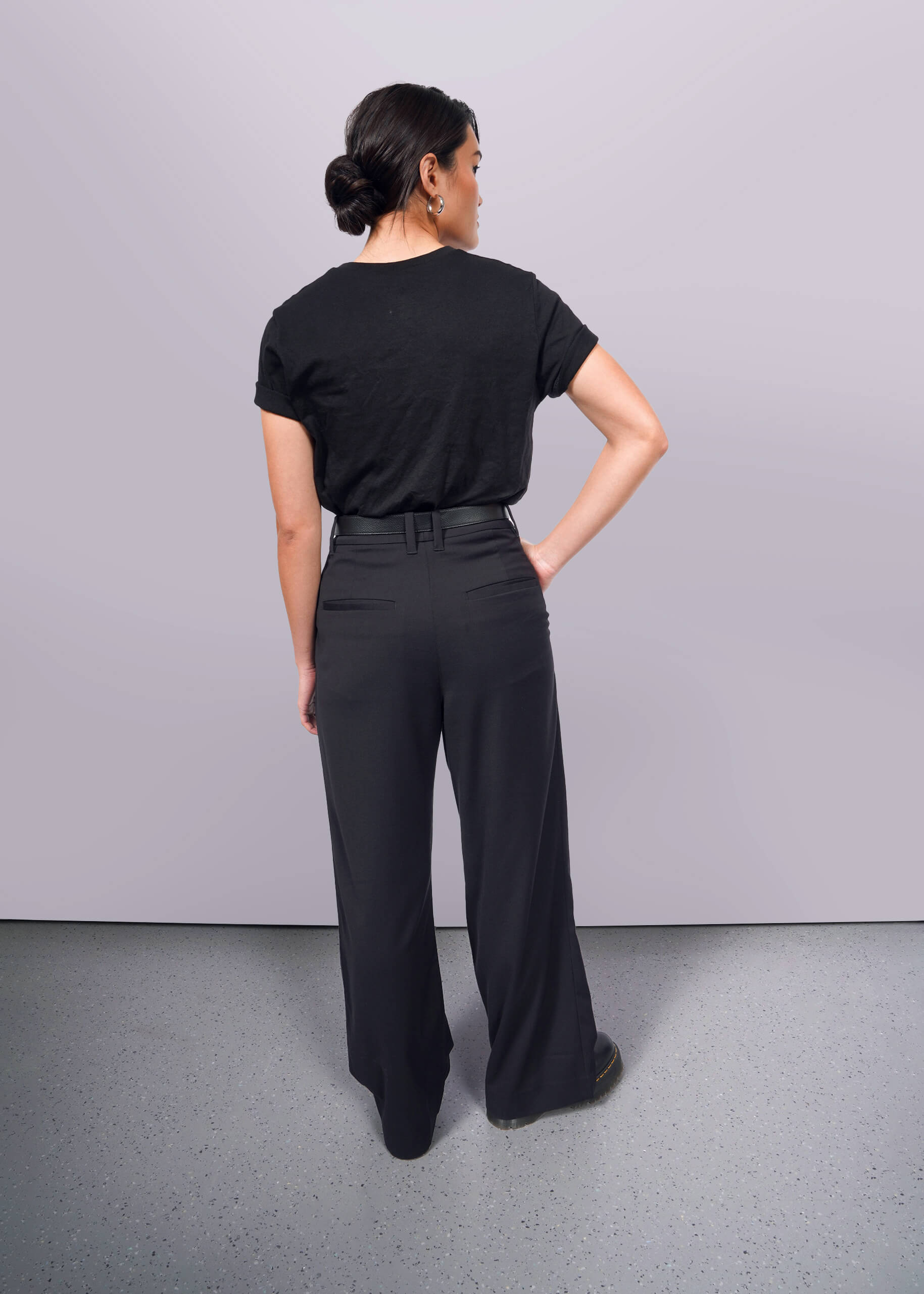 S=A person with dark hair in a bun stands facing away, wearing The Wild Feminist® black cotton unisex T-shirt and black wide-leg pants. One hand rests on their hip against a plain white background.