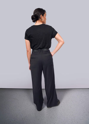 S=A person with dark hair in a bun stands facing away, wearing The Wild Feminist® black cotton unisex T-shirt and black wide-leg pants. One hand rests on their hip against a plain white background.