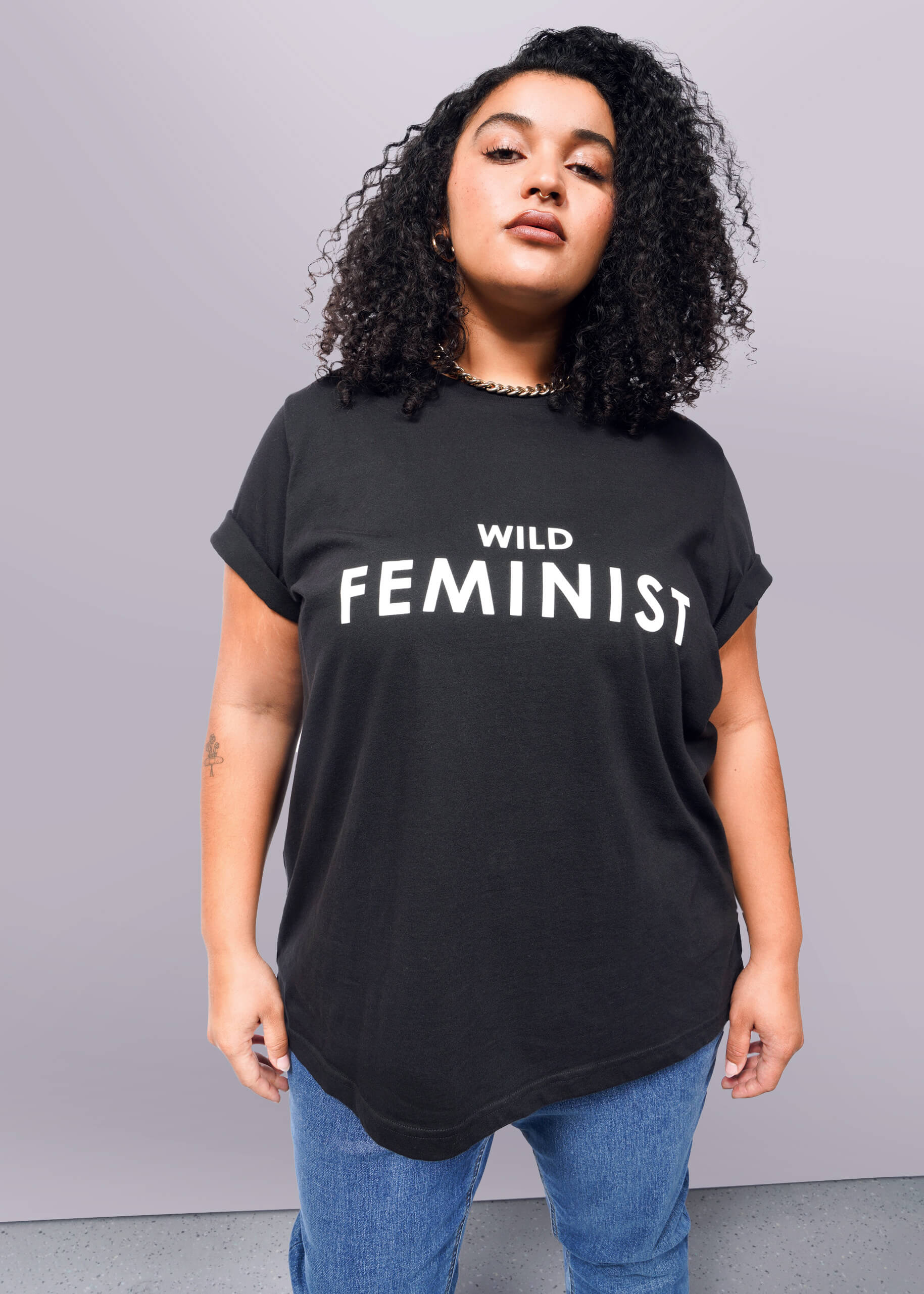 1X=Against a white background, a person with curly hair confidently wears The Wild Feminist® black tee, made of soft cotton and featuring bold white text. Paired with blue jeans, the unisex shirt reflects their expressive style.