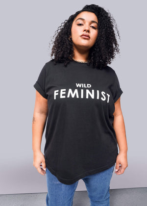 1X=Against a white background, a person with curly hair confidently wears The Wild Feminist® black tee, made of soft cotton and featuring bold white text. Paired with blue jeans, the unisex shirt reflects their expressive style.