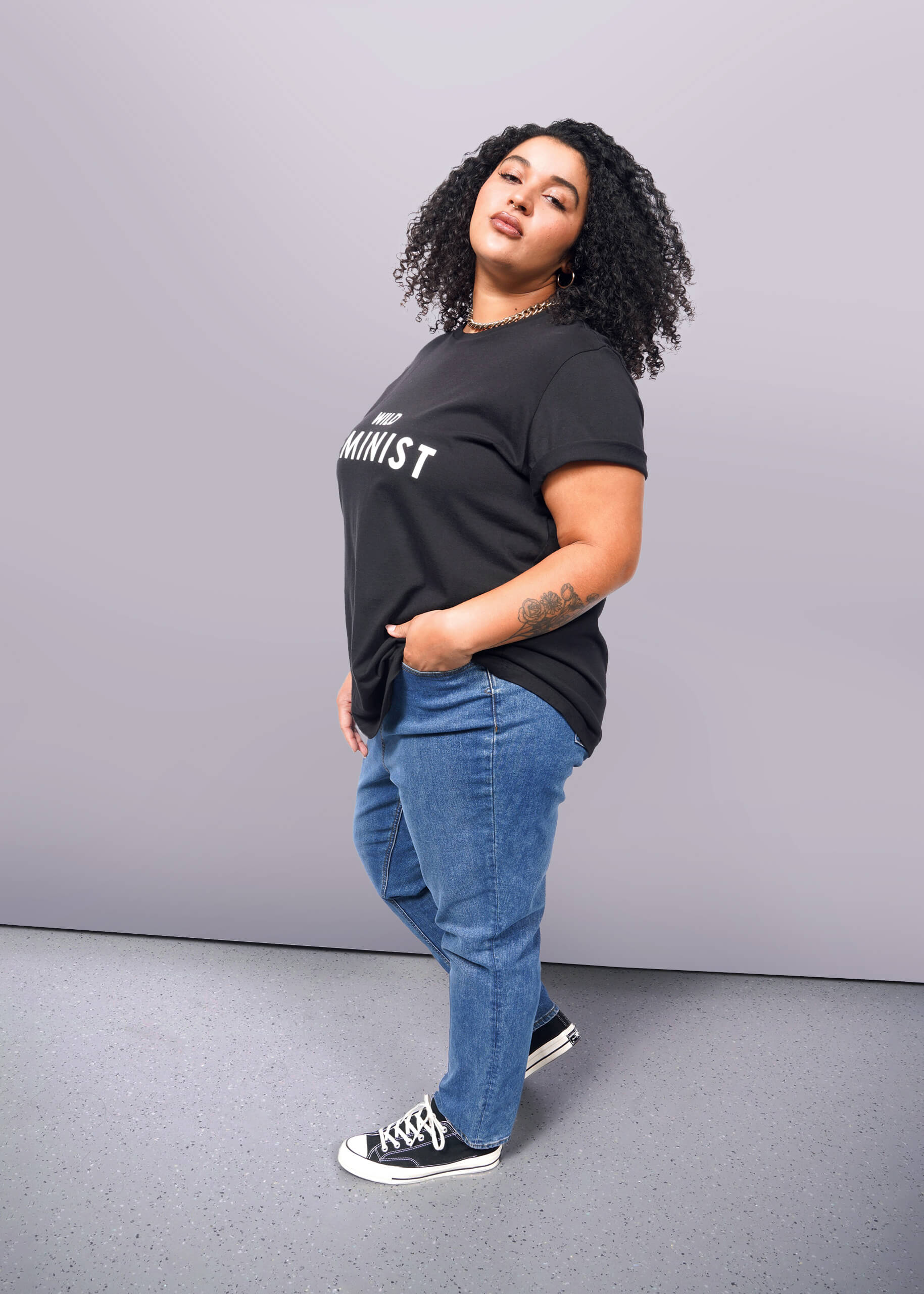 1X=A person with curly hair confidently wears The Wild Feminist® Tee, a black cotton unisex shirt with Feminist printed on it. Paired with blue jeans and black sneakers, they pose boldly against a plain white background.