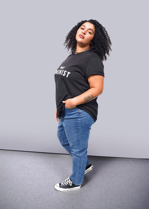 1X=A person with curly hair confidently wears The Wild Feminist® Tee, a black cotton unisex shirt with Feminist printed on it. Paired with blue jeans and black sneakers, they pose boldly against a plain white background.
