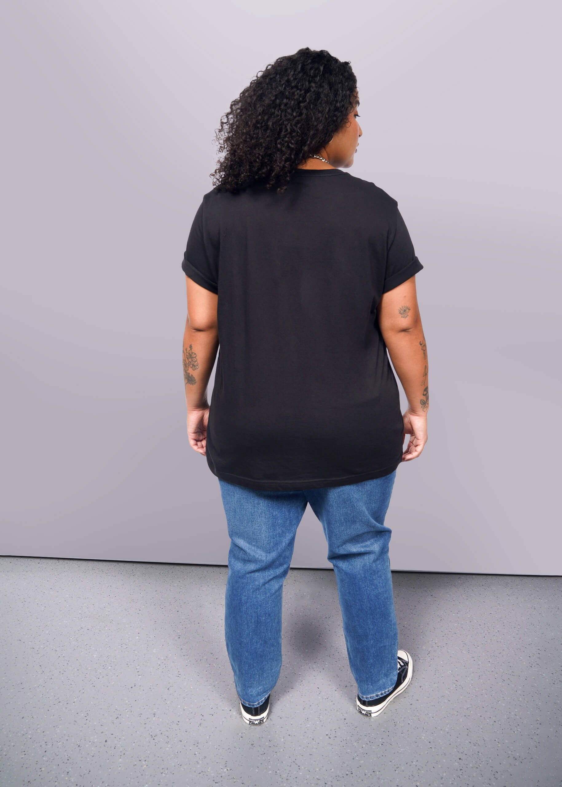 1X=A person with curly hair in a unisex black The Wild Feminist® Tee, blue jeans, and black sneakers stands on a gray floor, facing away from the camera. Tattoos adorn their arms.
