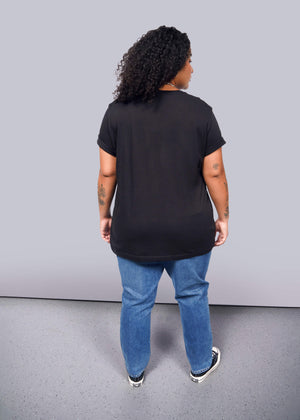 1X=A person with curly hair in a unisex black The Wild Feminist® Tee, blue jeans, and black sneakers stands on a gray floor, facing away from the camera. Tattoos adorn their arms.