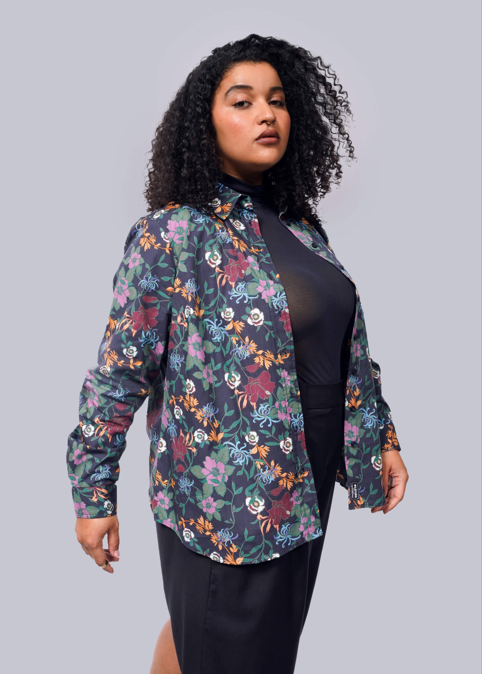 1X=Woman wearing a floral patterned shirt over a black top on a gray background