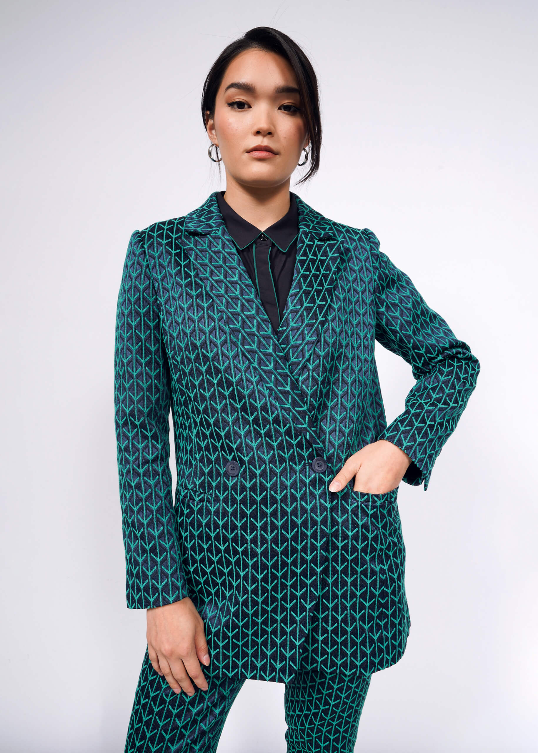 S= Standing confidently, a person wears The Empower Jacquard Double Breasted Blazer in teal with a geometric pattern over a black shirt, styled with hoop earrings and sleek hair. The woven jacquard fabric adds sophistication against the plain, light-colored background.