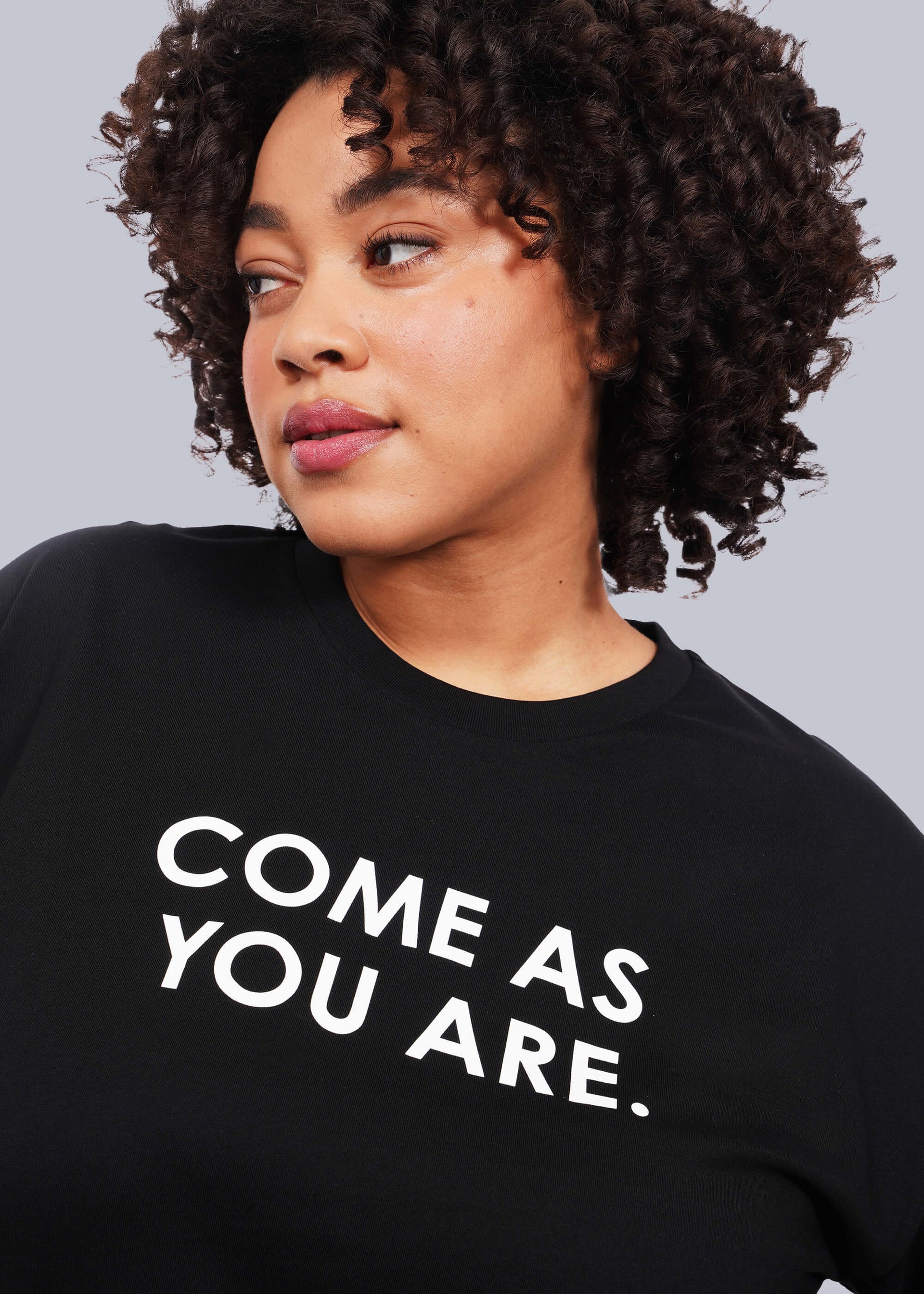 1X=Close up, chest up view of model with short curly hair wearing The Come As You Are Tee in black, which is a black relaxed fit tshirt with bold text across the chest that says "COME AS YOU ARE."