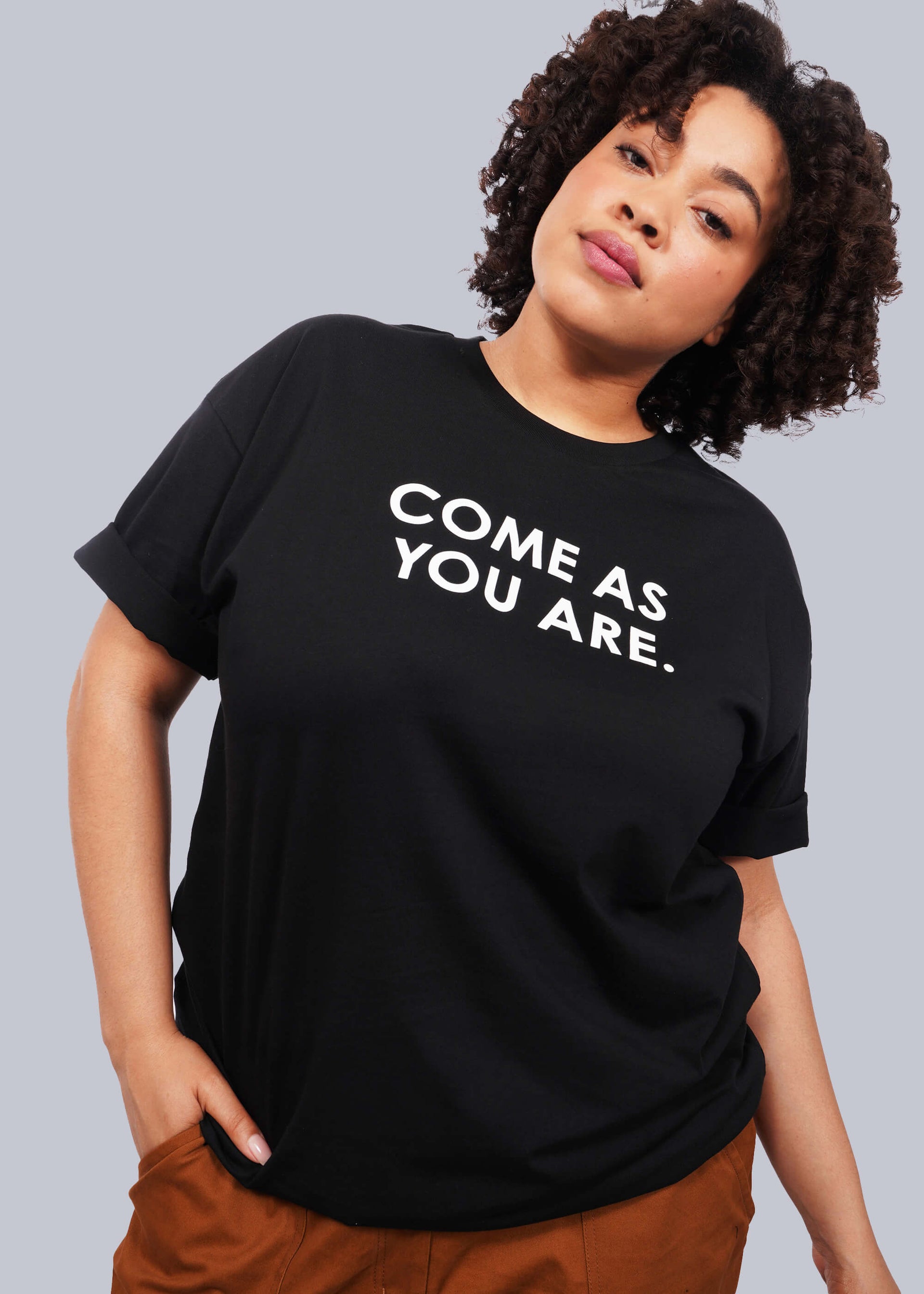 1X=Waist up image of model with short curly hair facing camera wearing The Come As You Are Tee in black, which is a black relaxed fit tshirt with bold text across the chest that says "COME AS YOU ARE."