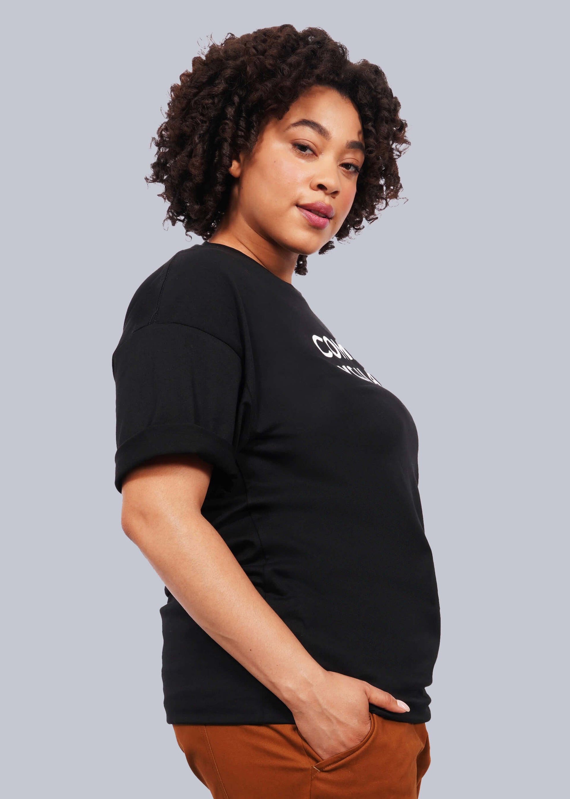 1X=Waist up side view image of model with short curly hair wearing The Come As You Are Tee in black, which is a black relaxed fit tshirt with bold text across the chest that says "COME AS YOU ARE." Model has hand in brown pants pocket.