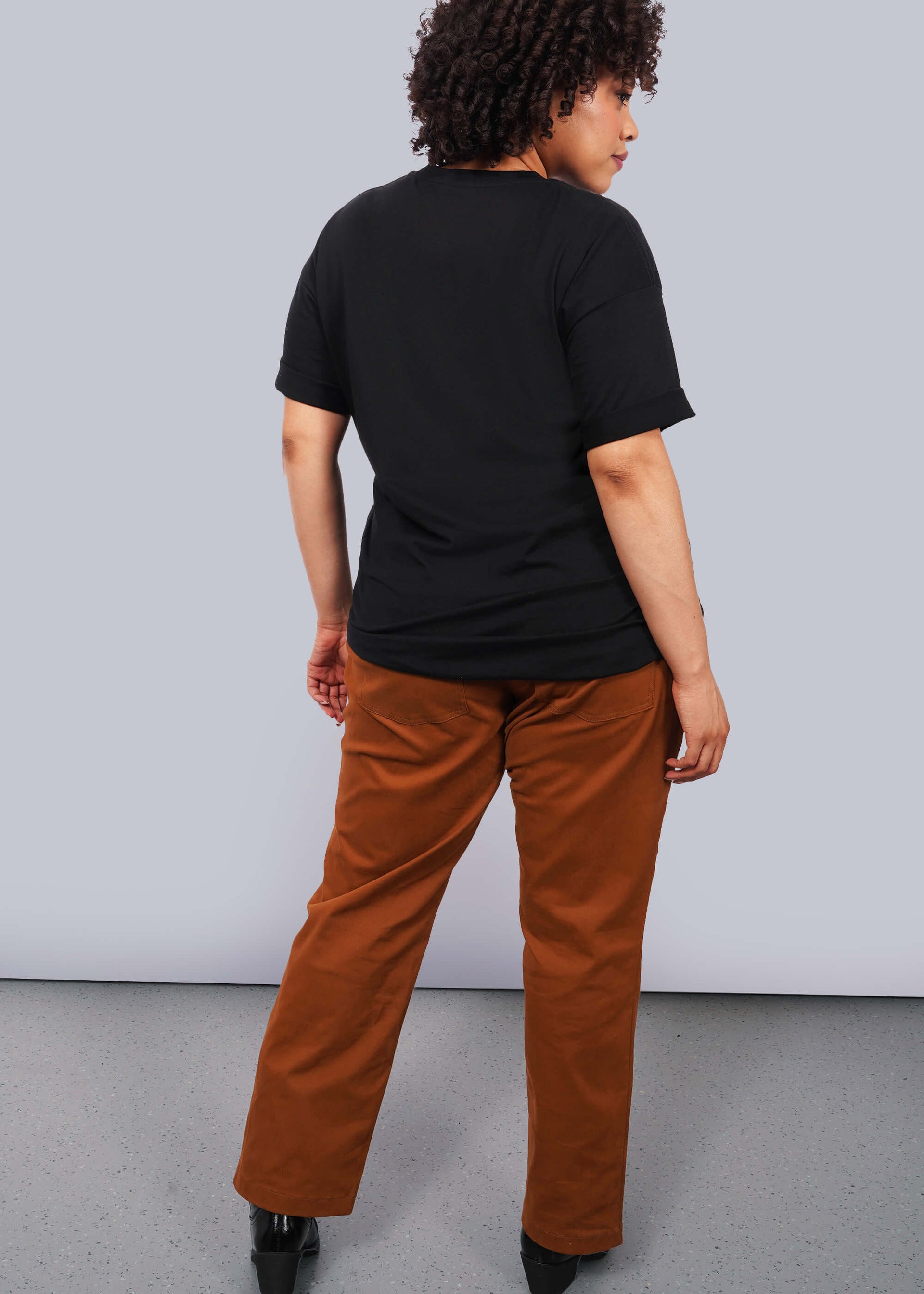1X=Full body back view image of model with short curly hair wearing brown pants and The Come As You Are Tee in black, which is a black relaxed fit tshirt with bold text across the chest that says "COME AS YOU ARE." The text is not visible in this image since this is a back view of the shirt.