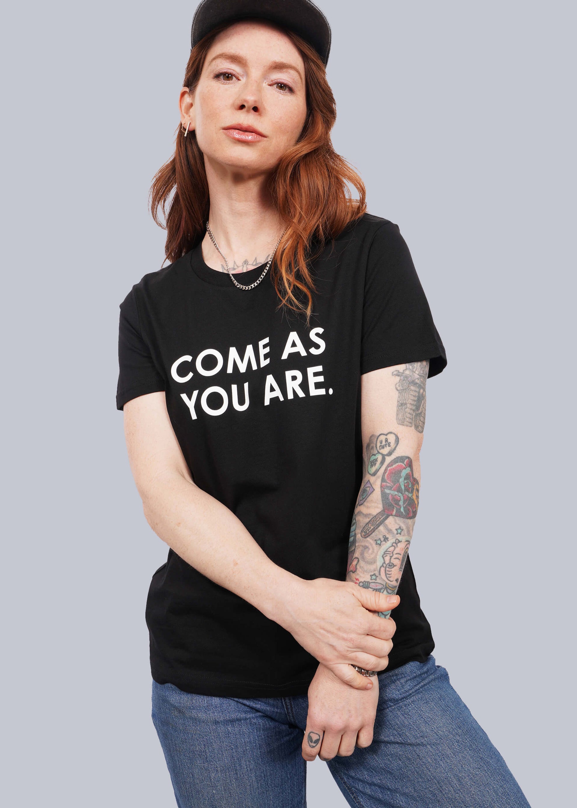 S=Model with wavy long red hair faces camera wearing a black hat and The Come As You Are Tee in Black, which is a black relaxed fit tshirt with bold white text across the chest that says "COME AS YOU ARE."