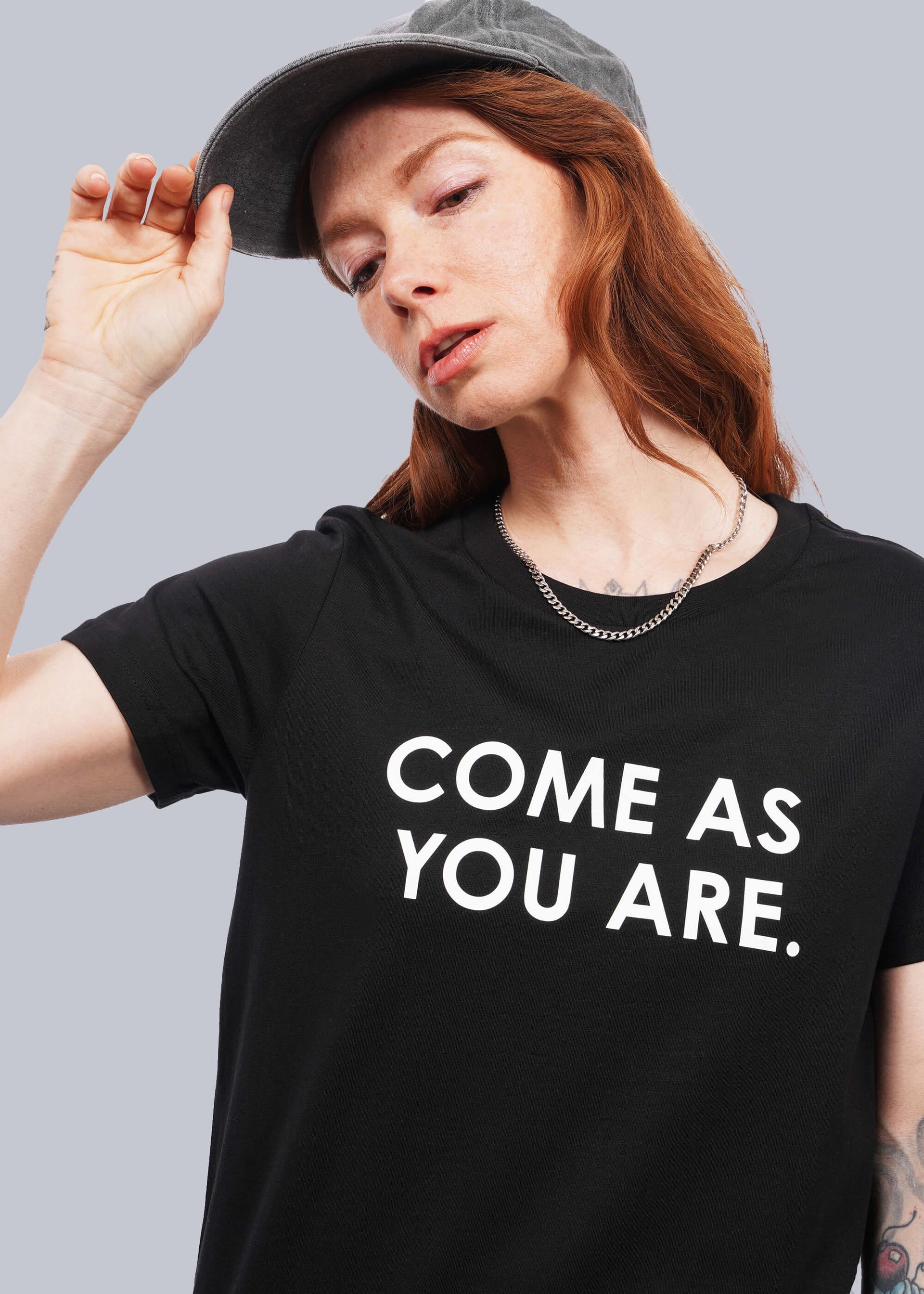 S=Close up of model with wavy long red hair facing camera wearing a cap and The Come As You Are Tee in Black, which is a black relaxed fit tshirt with bold white text across the chest that says "COME AS YOU ARE."