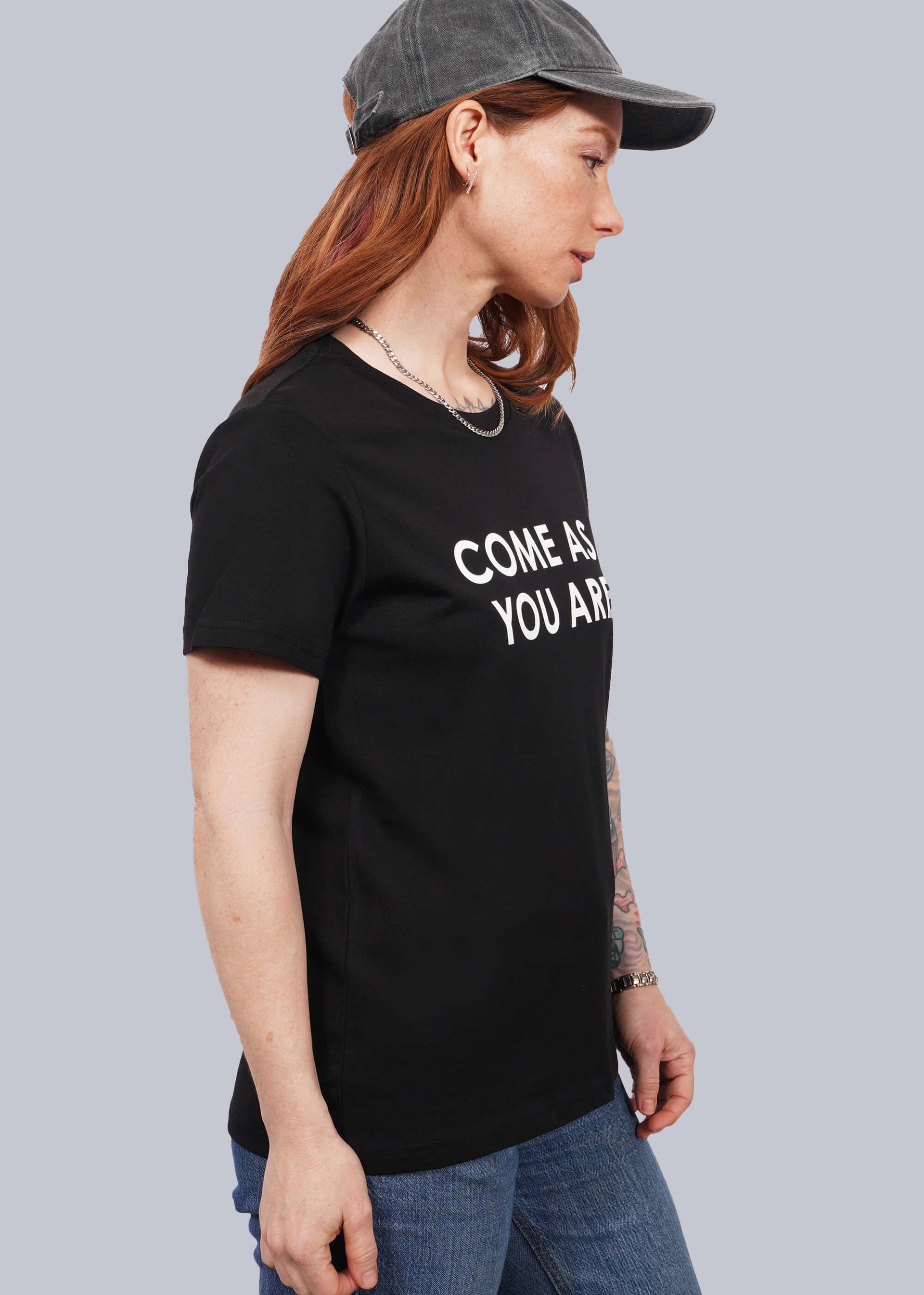 S=Waist up side view of model with wavy long red hair wearing a cap, jeans, and The Come As You Are Tee in Black, which is a black relaxed fit tshirt with bold white text across the chest that says "COME AS YOU ARE."