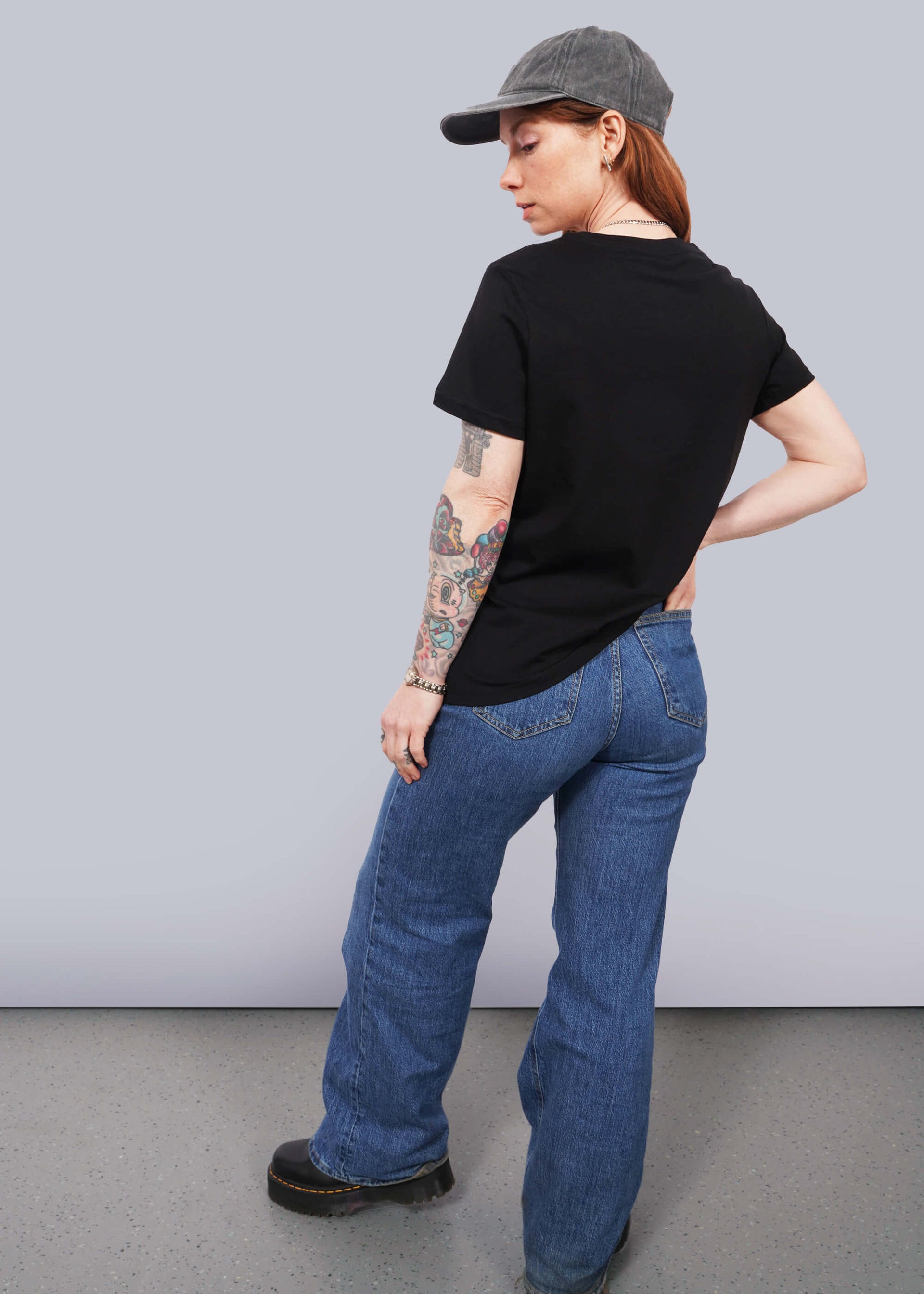 S=Full body back view of model with wavy long red hair wearing a cap, jeans, and The Come As You Are Tee in Black, which is a black relaxed fit tshirt with bold white text across the chest that says "COME AS YOU ARE." Text is not visible in this view since it shows the back of the shirt.