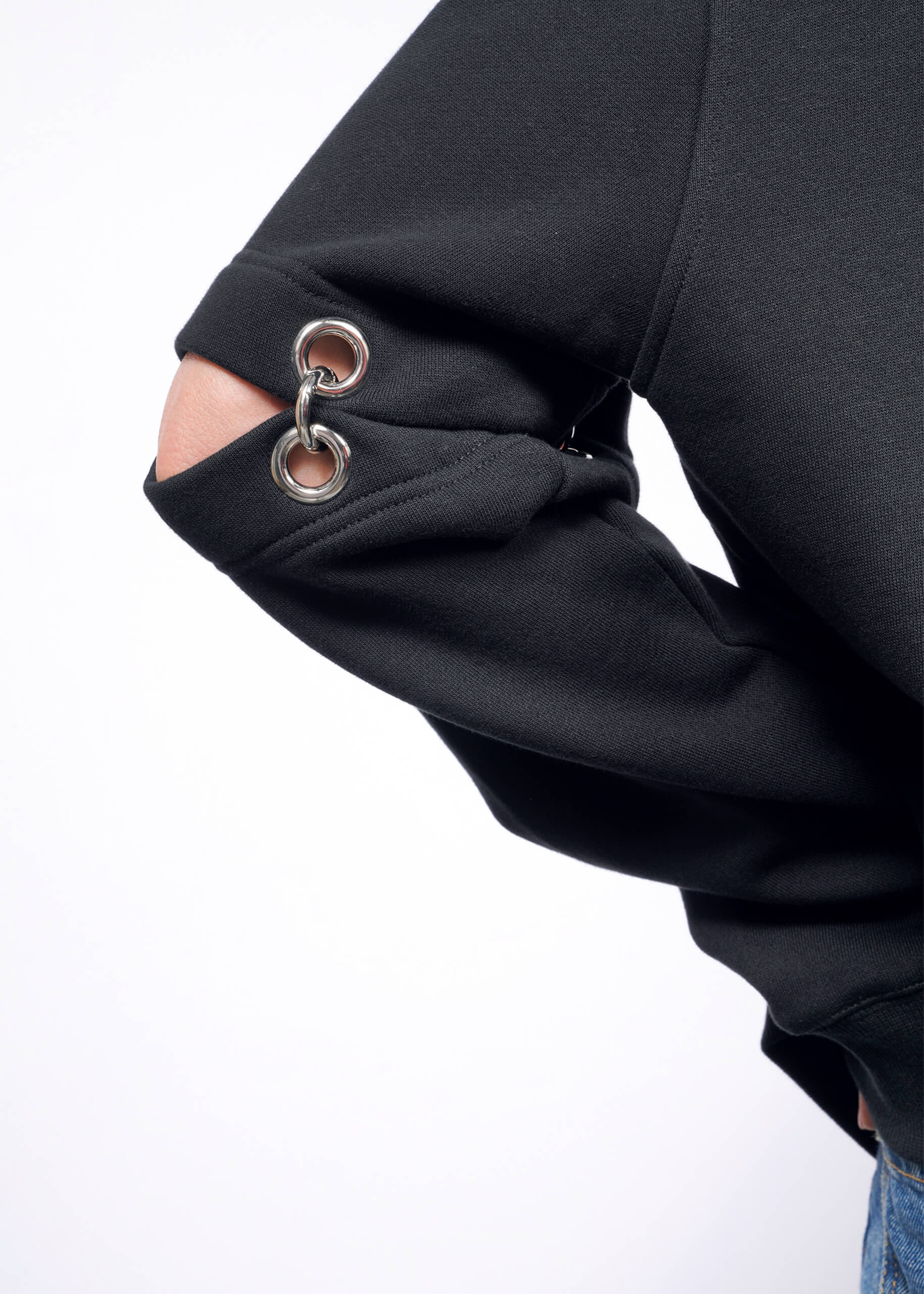 S=A person showcases the Debbie Harry X Wildfang Convertible Sweatshirt, a black jacket with unique removable sleeve cut-outs featuring metal grommets, as their hand rests on their hip against a white background.