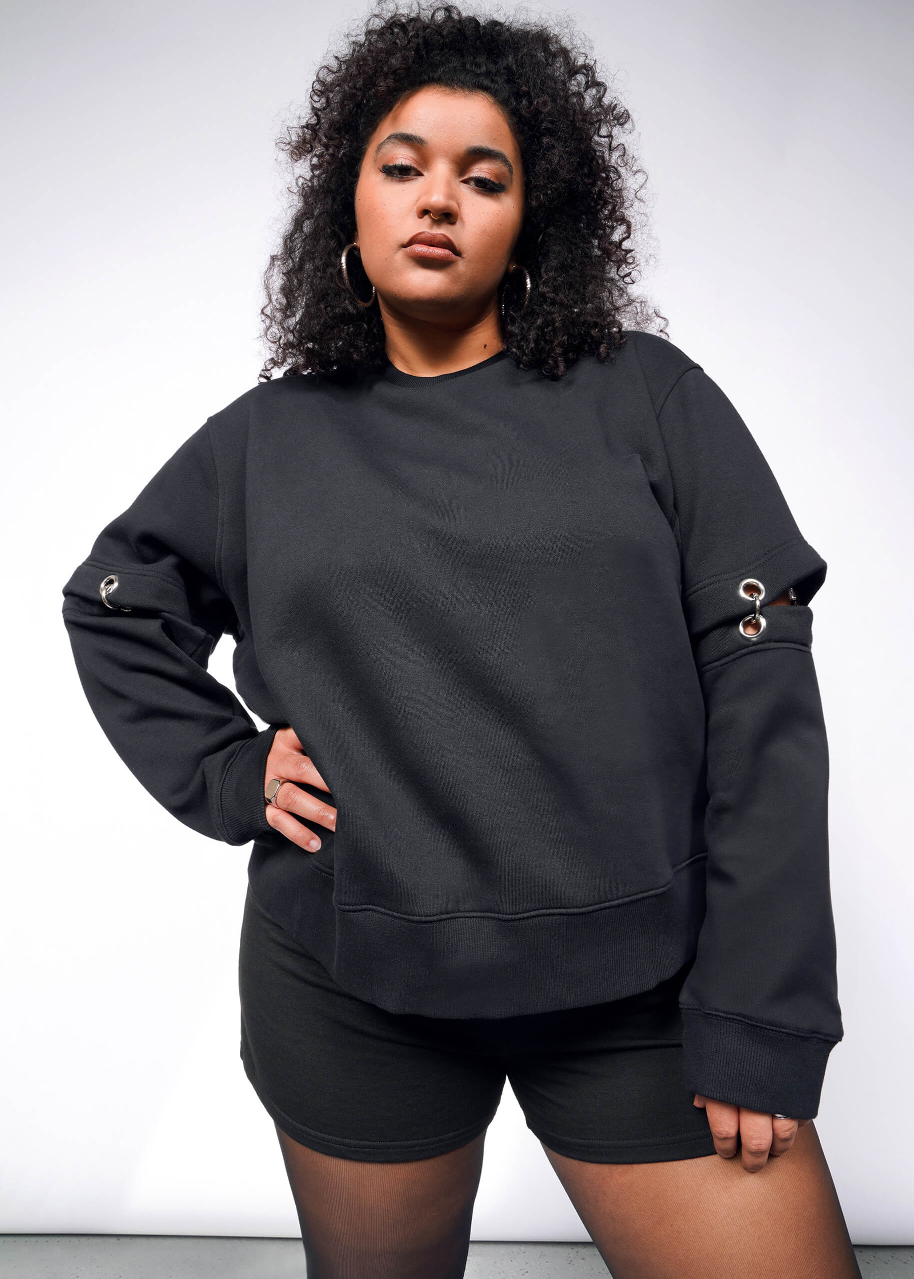 1X= Someone with curly hair poses confidently against a white backdrop in the Debbie Harry X Wildfang Convertible Sweatshirt, showcasing its metal eyelet details and removable sleeves, paired with matching black shorts. They convey a strong, self-assured vibe complemented by hoop earrings.