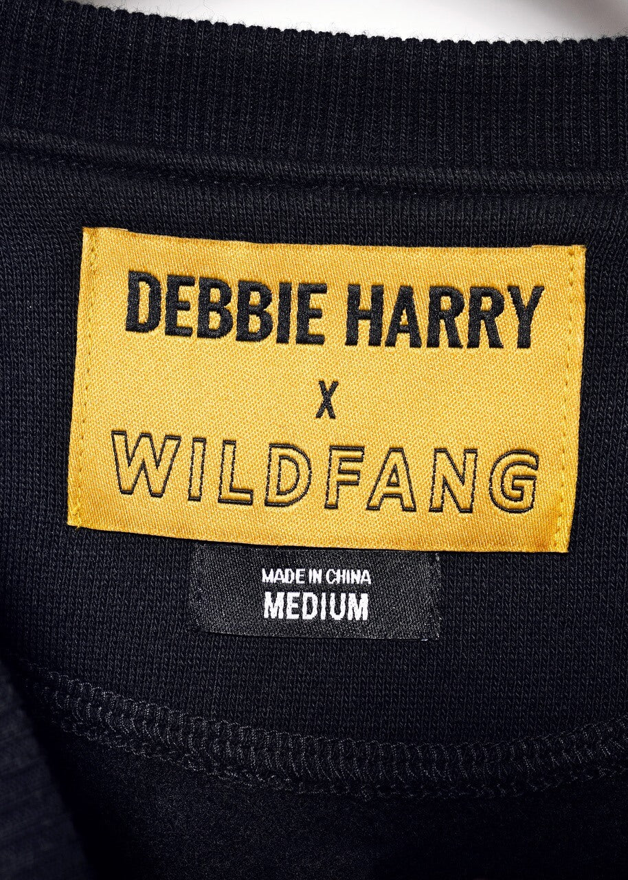 S=Close-up of clothing labels on a black Debbie Harry X Wildfang Convertible Sweatshirt. The yellow label says DEBBIE HARRY x WILDFANG, and a smaller black label reads MADE IN CHINA MEDIUM, suggesting features like removable sleeves.