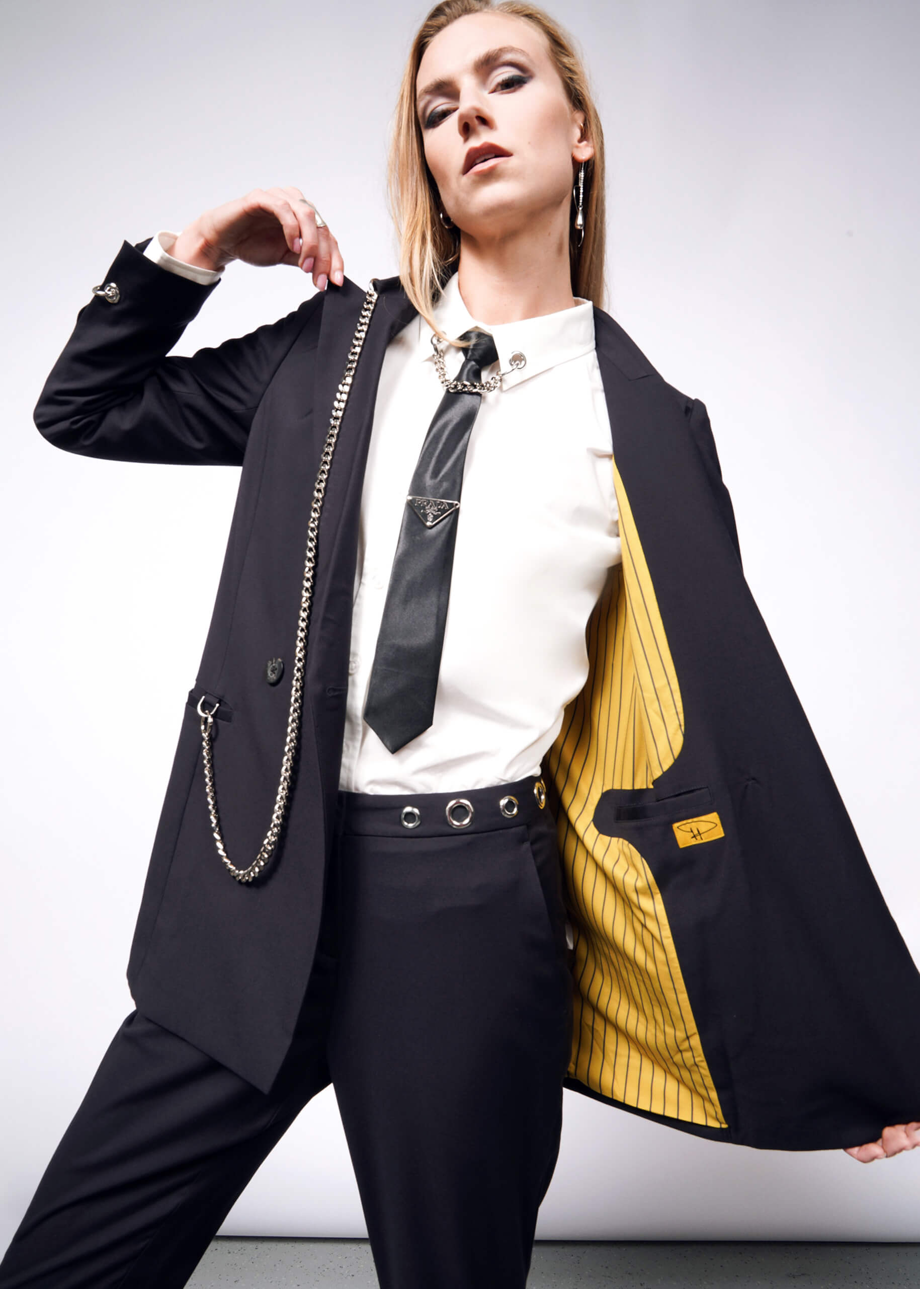 S=A stylish person poses confidently in a black suit with a white shirt and tie, showcasing the Debbie Harry X Wildfang Double Breasted Blazers striking yellow lining. The outfit is perfectly accessorized with chains and exquisite metal detailing.