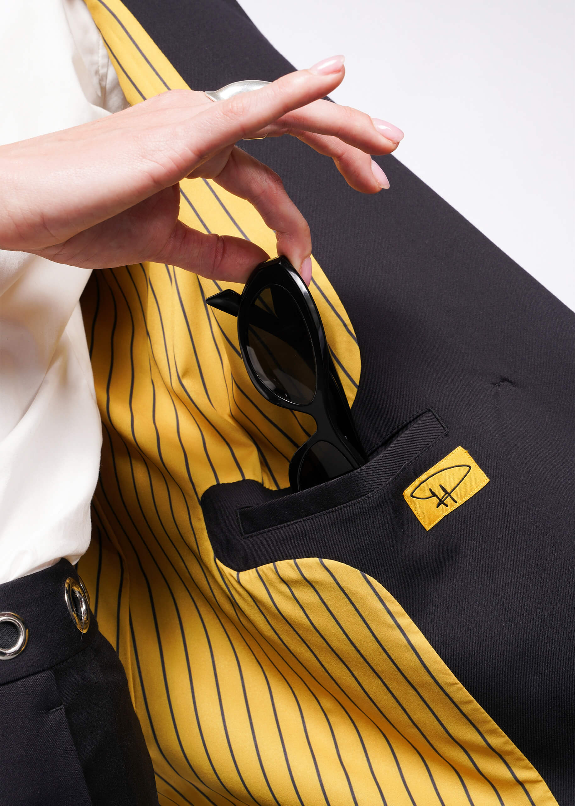 S=Black sunglasses are slipped into the inner pocket of a Debbie Harry X Wildfang Double Breasted Blazer, with yellow and black striped lining featuring an eyeglass frame logo. A person pairs it with a white shirt and dark pants adorned with metal detailing.