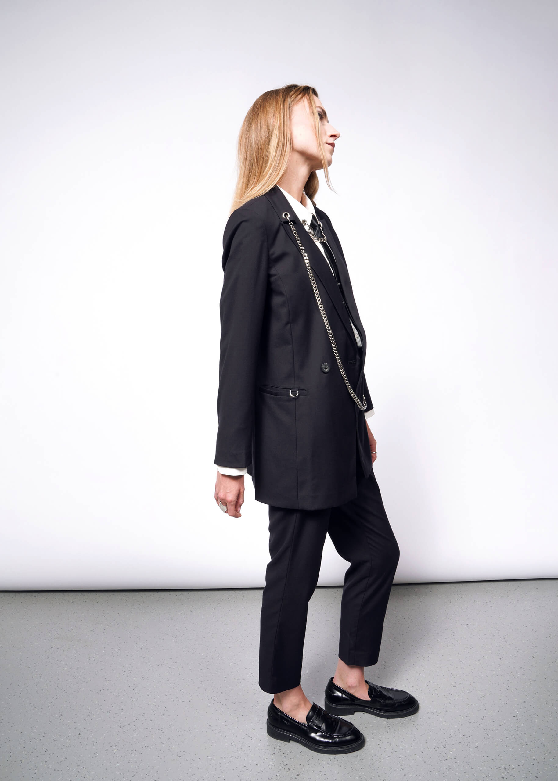 S=A person with long hair in profile against a plain background wears a Debbie Harry X Wildfang Double Breasted Blazer with metal detailing. They pair it with shiny black loafers, looking upward, their hands relaxed at their sides.