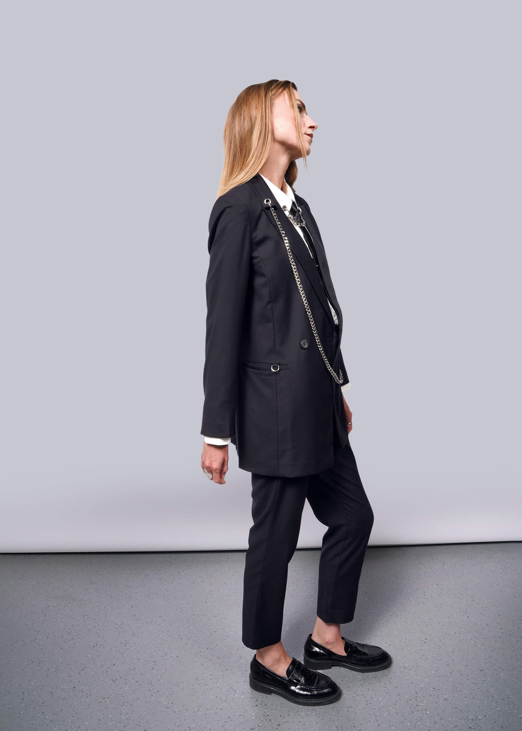 S=A person with long hair in profile against a plain background wears a Debbie Harry X Wildfang Double Breasted Blazer with metal detailing. They pair it with shiny black loafers, looking upward, their hands relaxed at their sides.
