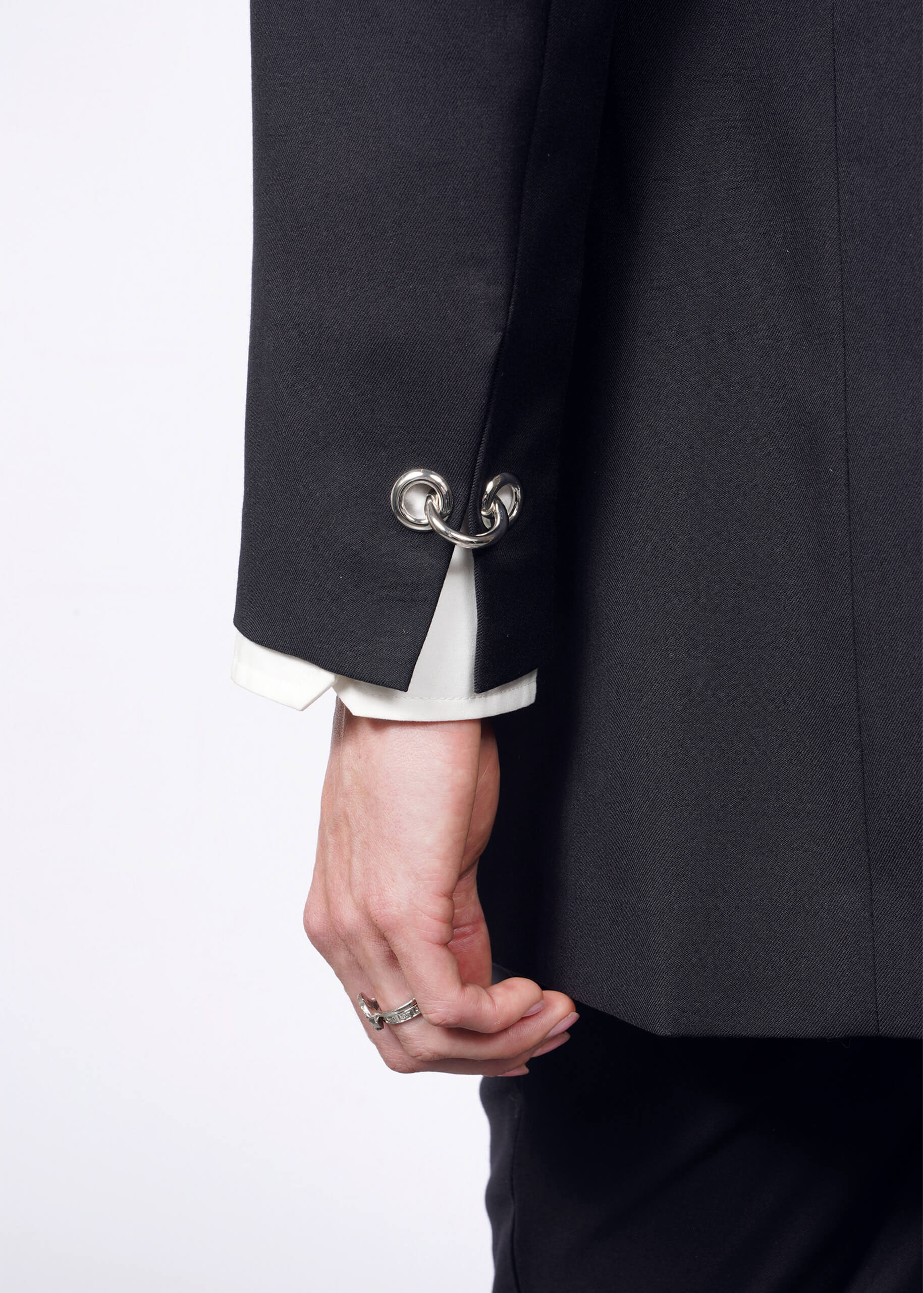 S= A person wears the sleek black Debbie Harry X Wildfang Double Breasted Blazer featuring metal hoop cufflinks while holding the lapel. A white shirt sleeve shows, paired with a silver ring on their middle finger, contrasting against a white background and highlighting striking metal details.