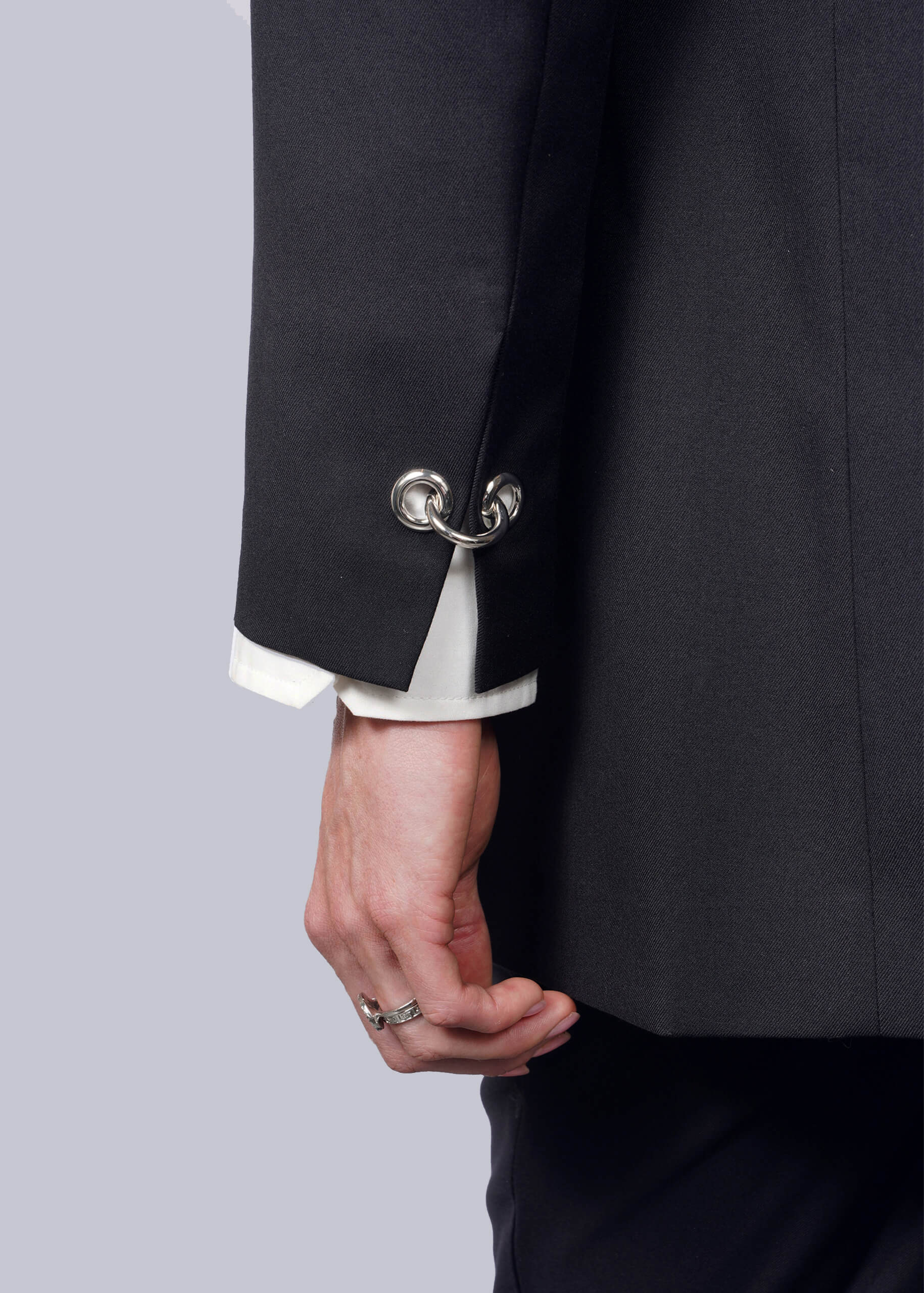 S= A person wears the sleek black Debbie Harry X Wildfang Double Breasted Blazer featuring metal hoop cufflinks while holding the lapel. A white shirt sleeve shows, paired with a silver ring on their middle finger, contrasting against a white background and highlighting striking metal details.