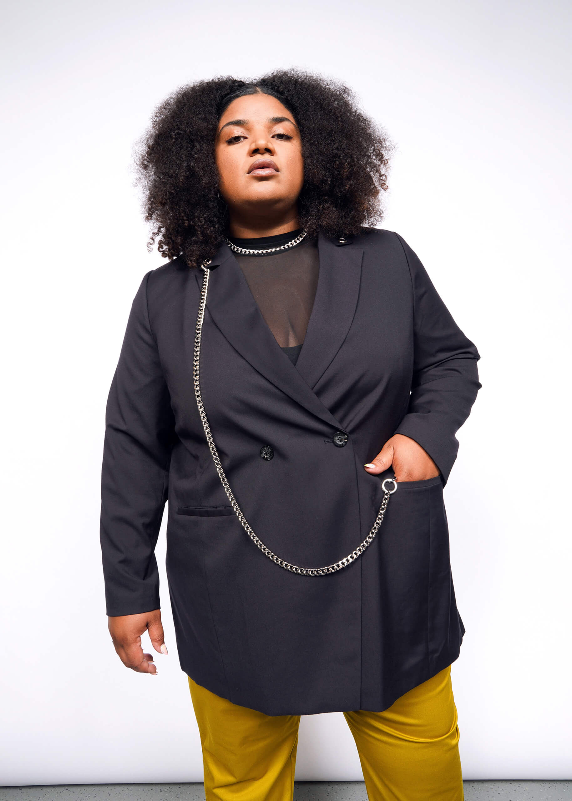 1X= A person with curly hair confidently poses in a stylish Debbie Harry X Wildfang Double Breasted Blazer, featuring intricate metal detailing over a sheer black top and vibrant yellow pants, set against a plain white background.