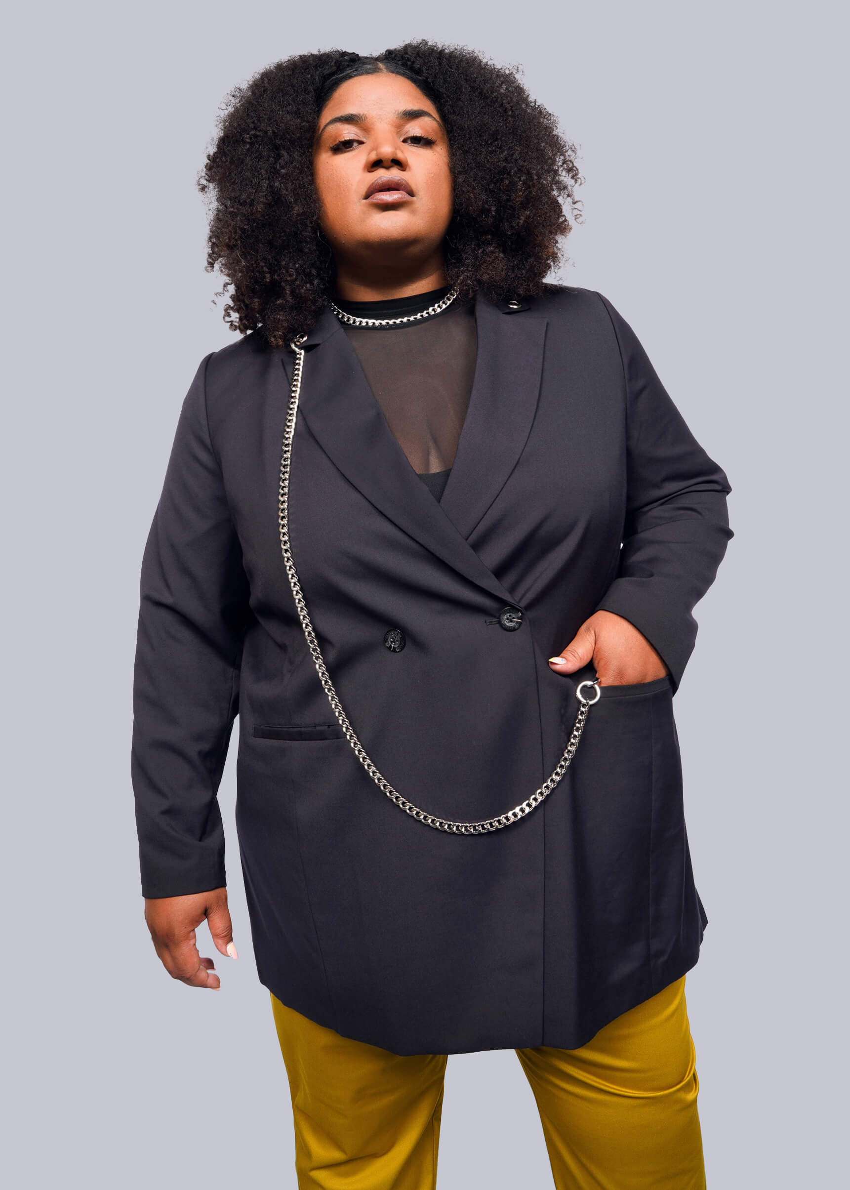 1X= A person with curly hair confidently poses in a stylish Debbie Harry X Wildfang Double Breasted Blazer, featuring intricate metal detailing over a sheer black top and vibrant yellow pants, set against a plain white background.
