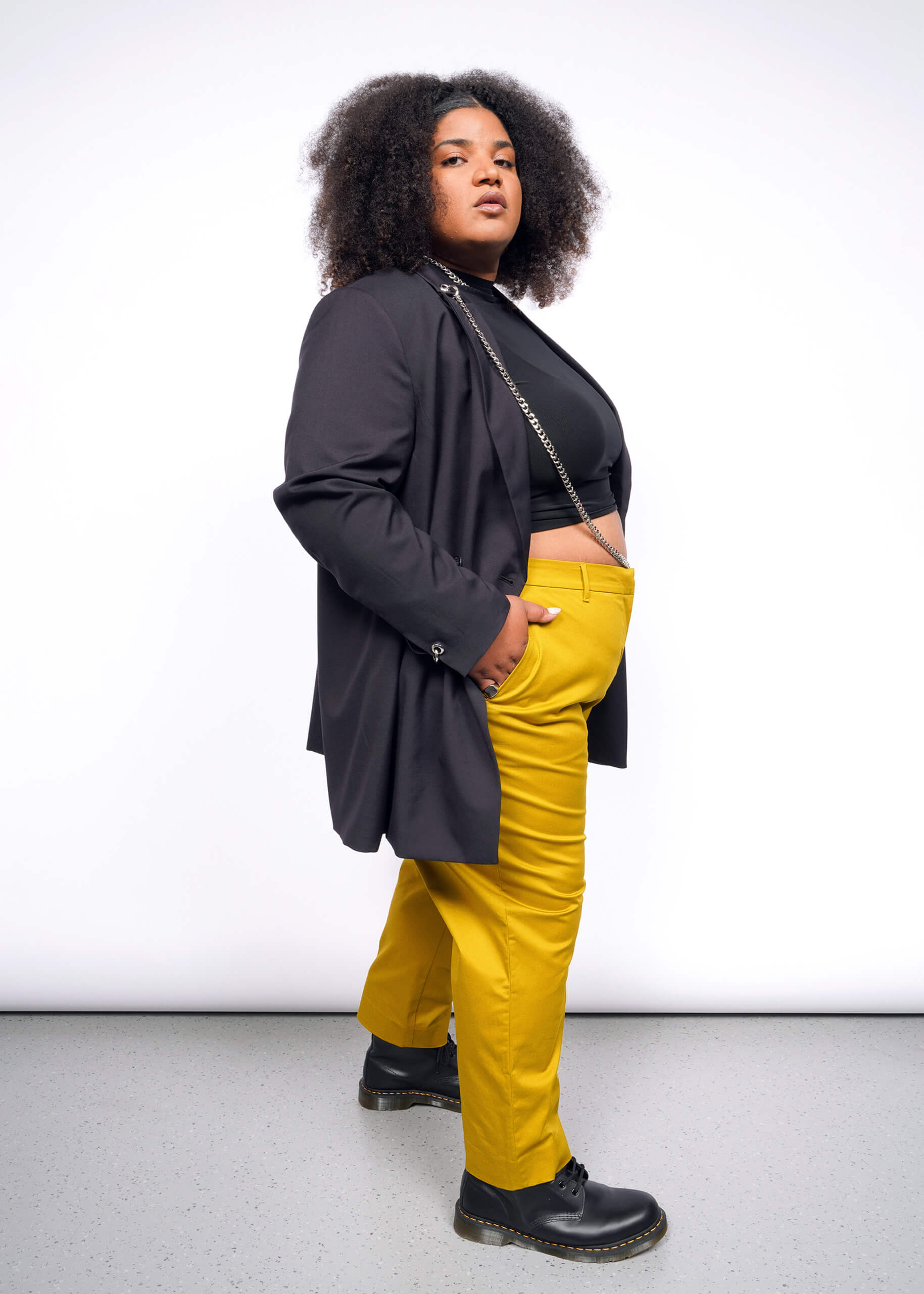 1X=A person with curly hair confidently poses, wearing a Debbie Harry X Wildfang Double Breasted Blazer over a black top and bright yellow pants. The outfit, featuring subtle metal detailing, complements their stance on a grey floor against a plain white backdrop.