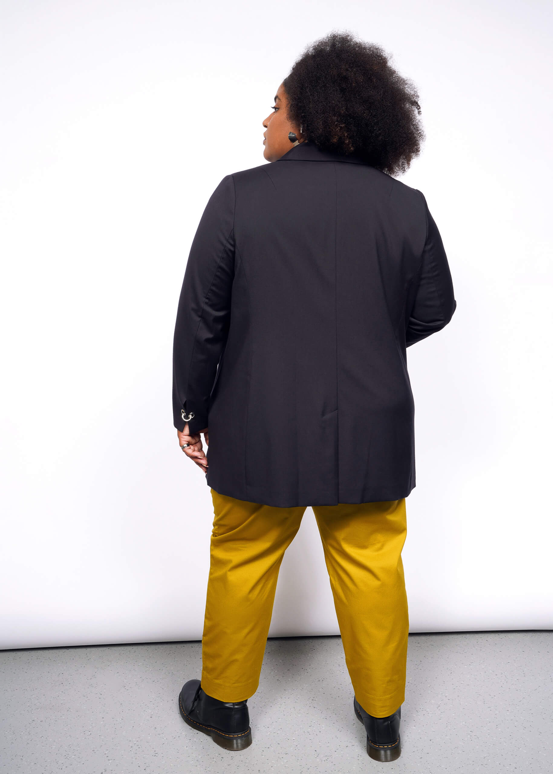 1X= Showcasing a Debbie Harry X Wildfang Double Breasted Blazer with subtle metal details, the person pairs it stylishly with mustard yellow pants and black shoes. Their curly hair cascades down their back as they stand against a pristine white background on a light gray floor.