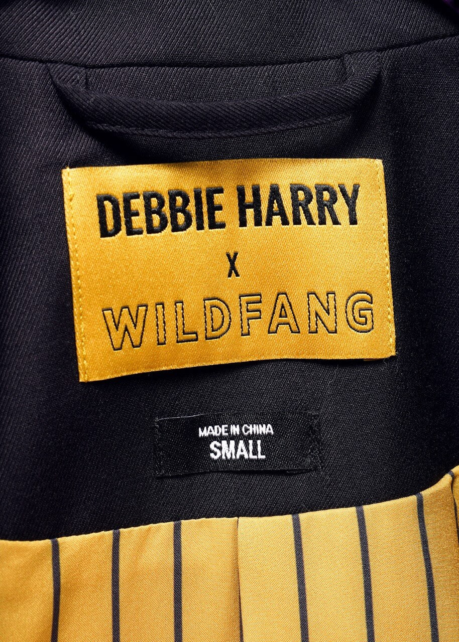 S=Close-up of a DEBBIE HARRY X WILDFANG label inside the Debbie Harry X Wildfang Double Breasted Blazer. The label is in bold black text on yellow, stating MADE IN CHINA SMALL. The blazer has yellow and black vertical stripes with metal detailing.