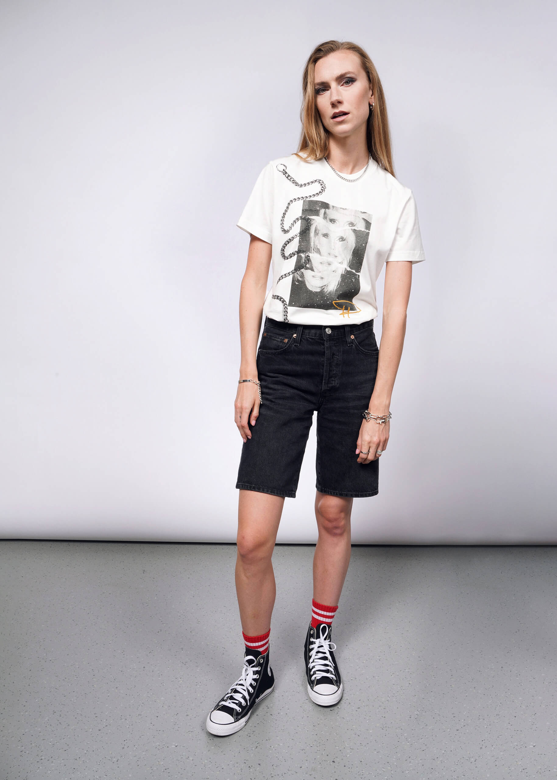 S=Wearing the Debbie Harry X Wildfang Graphic Tee with black knee-length shorts, black Converse sneakers, and red striped socks, a long-haired person exudes punk style reminiscent of Debbie Harry. They stand relaxed on a gray speckled floor against a neutral background.