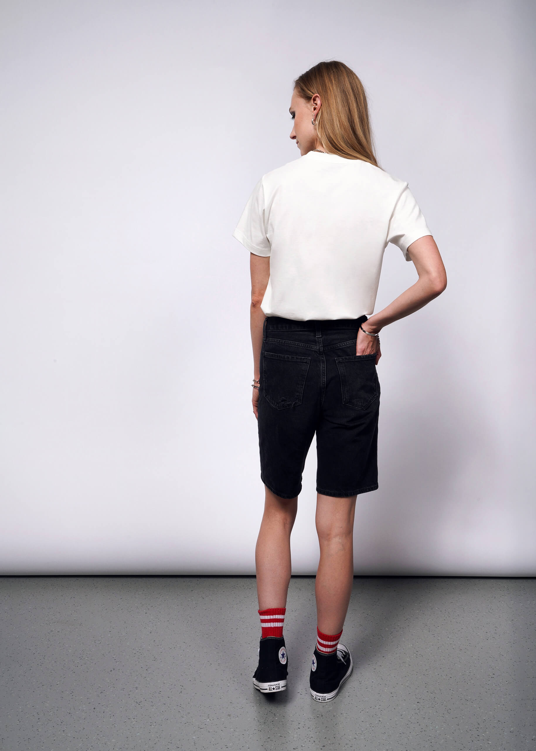 S=With long hair reminiscent of Debbie Harrys style, a person is seen from the back in a minimalist room with light grey floors and walls. Theyre wearing a Debbie Harry X Wildfang Graphic Tee, black denim shorts, black high-top sneakers, and red striped socks for a punk aesthetic.