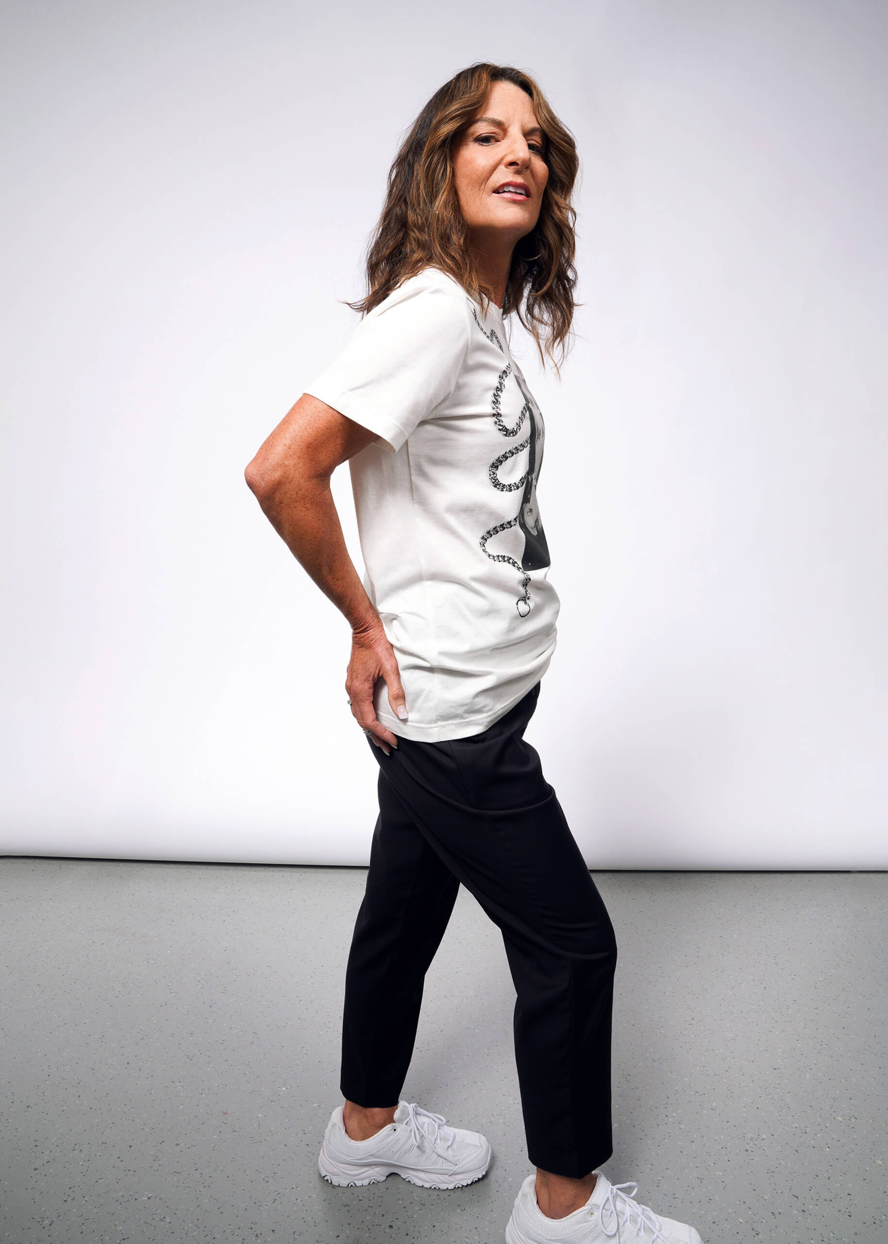 S=A person with long hair stands confidently in a Debbie Harry X Wildfang Graphic Tee and black pants. Channeling a punk aesthetic, they wear white sneakers, reminiscent of Debbie Harrys iconic style, posed against a plain gray background.