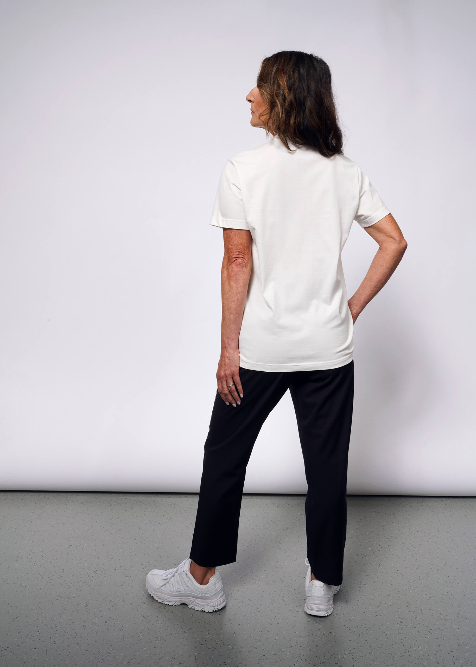 S= Facing away in a punk style, a woman wears the Debbie Harry X Wildfang Graphic Tee with black pants and white sneakers. Her right hand rests on her hip as she gazes slightly left, creating a modern-day Debbie Harry vibe against the plain light gray background.